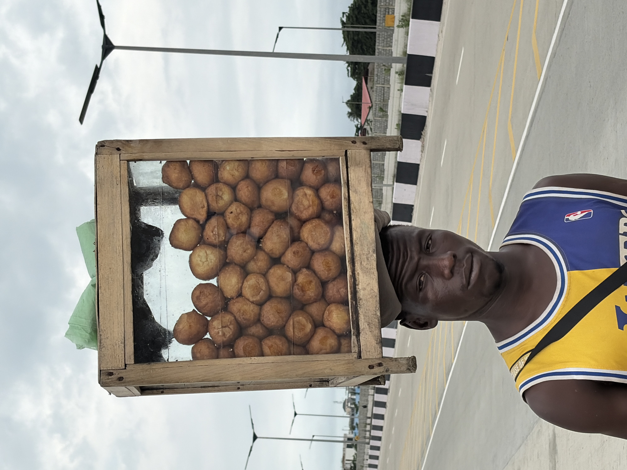 The image shows a man standing on an empty road, possibly in Igboefon, Nigeria. He is wearing a blue and yellow basketball jersey and carrying a wooden and glass display box filled with fried dough balls on his head. The box is propped up by a light-colored cap worn by the man. The background features a cloudy sky, streetlights, and a barrier along the side of the road. The man is looking directly at the camera with a neutral expression.