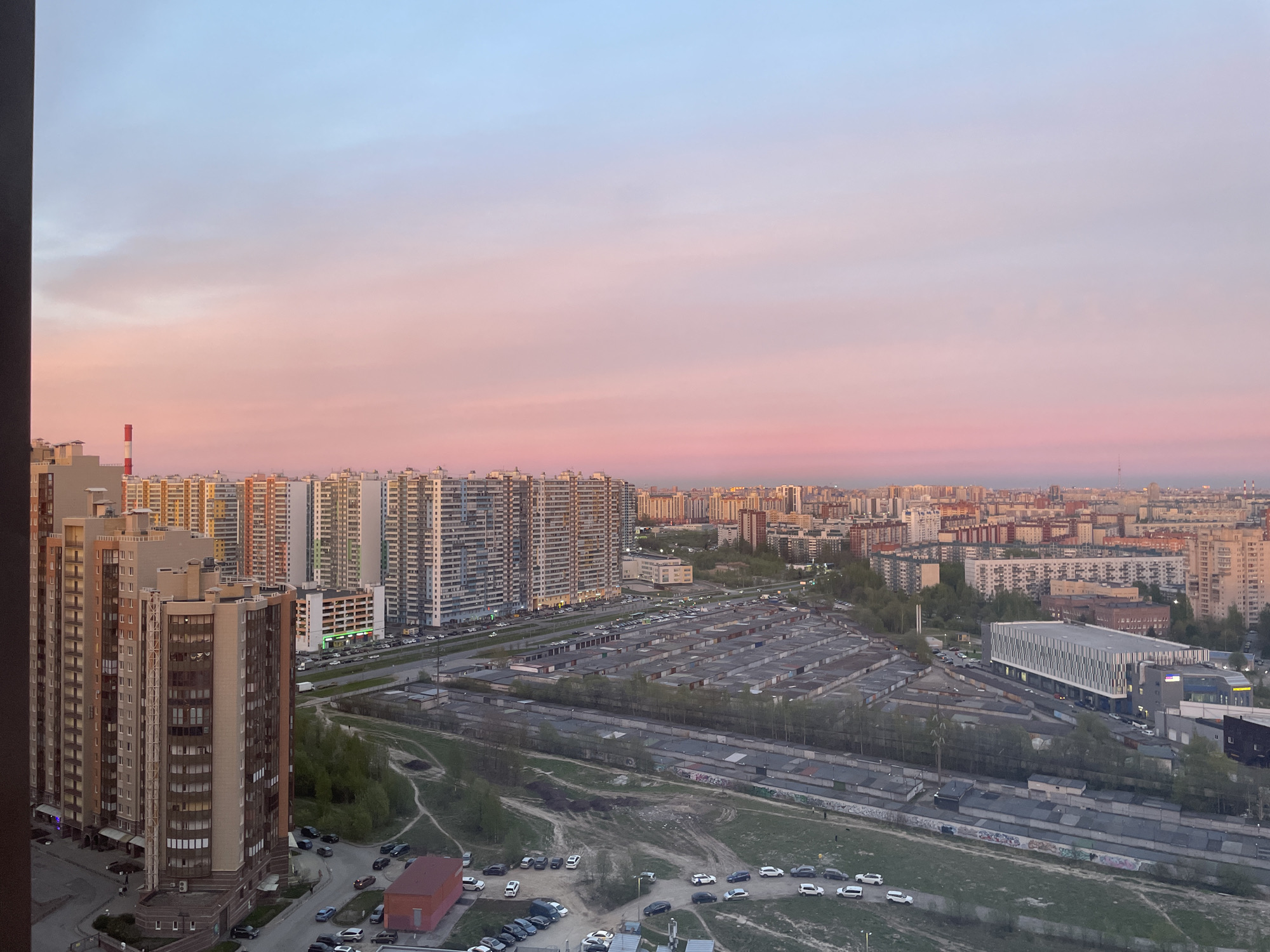 The image shows a view of a cityscape at dusk, possibly in Saint Petersburg, Russia. The sky is colored in shades of blue, pink, and white, indicating either sunrise or sunset. The cityscape includes several high-rise apartment buildings of varying colors and architectural styles. A large number of garage boxes are visible in the middle ground, stretching out across a considerable area. Several rows of parked cars are near them. In the foreground, more parked cars are visible. The overall environment suggests an urban area with a mix of residential buildings and infrastructure for transportation and storage. A single red and white chimney is visible on the left side of the photograph.