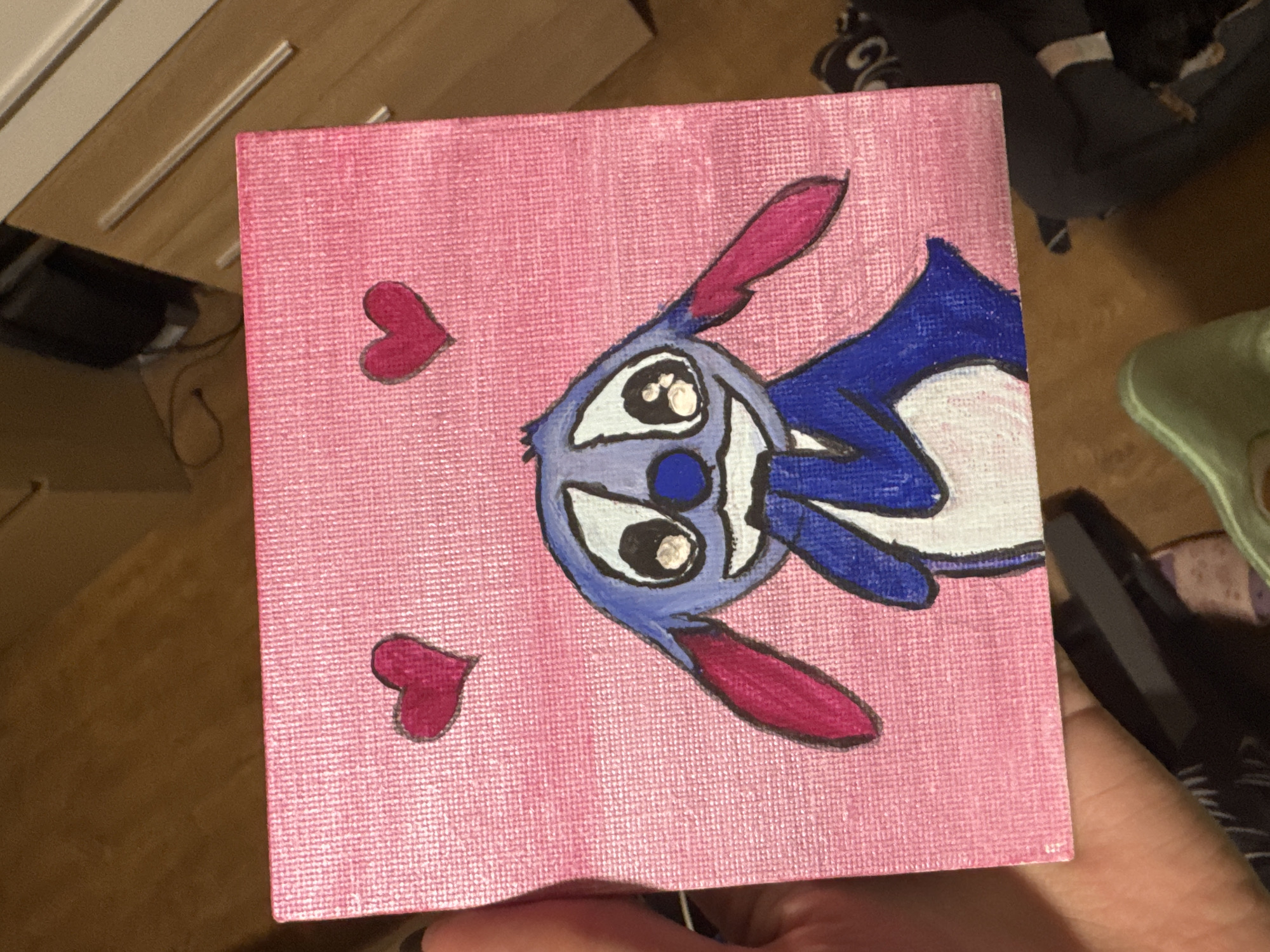 The image shows a square canvas painting with a pink background. It features a cartoon character resembling Angel from Lilo & Stitch, colored with shades of blue, white, and pink. Two small red hearts are painted on the canvas, one above and one below the character. The canvas is being held in a person's hand. The background includes a wooden surface, possibly a table or floor, and glimpses of other objects suggesting an indoor setting. There is no indication of the city of Sofia, Bulgaria.