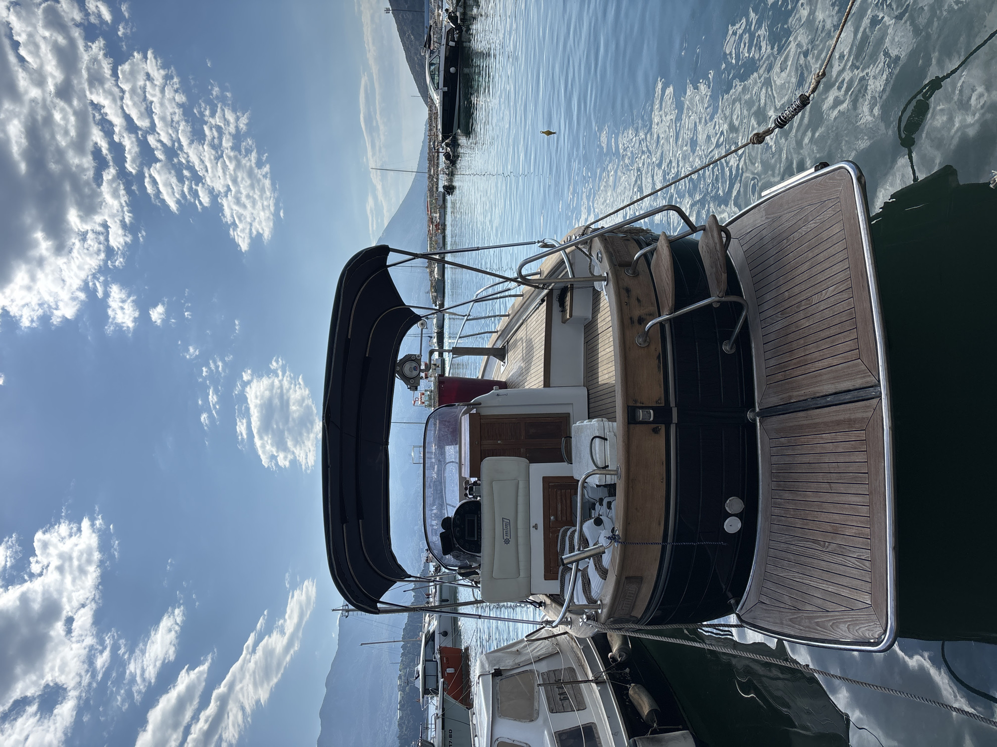 The image shows a boat docked at a marina in Budva, Montenegro. The boat is positioned sideways in the frame, with its stern featuring teak decking, metal railings, and a dark hull. A canvas awning is partially visible overhead. The boat's interior includes seating, a console, and wood cabinetry. Other boats are visible in the background, along with a mountain range and clear blue sky with scattered clouds. The water has ripples and reflections. The location is identified as Budva, Montenegro.