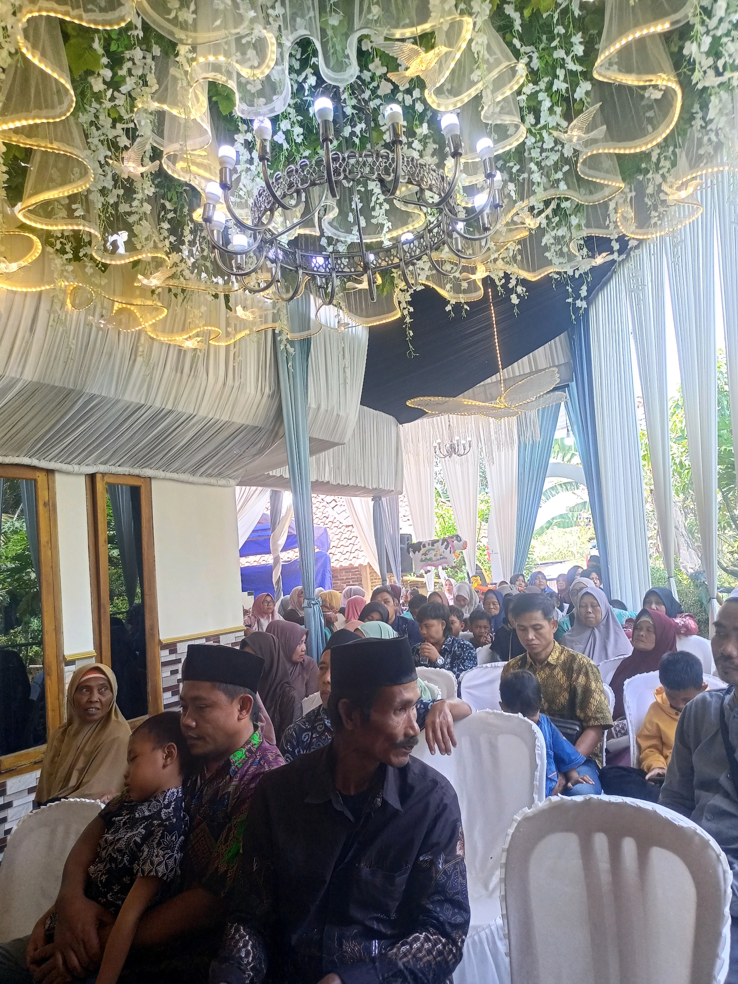 The image depicts an outdoor gathering, possibly a wedding or celebration, in Badamita, Indonesia. A large group of people, predominantly Muslim, are seated in rows on white chairs, attending an event. The foreground features several individuals, including a man in a black shirt holding a sleeping child. The backdrop is decorated with white drapes, artificial flowers, and a chandelier, creating an elegant ambiance. Natural light filters in from the right, illuminating the scene. The presence of traditional Indonesian attire and the architecture suggest a cultural event.