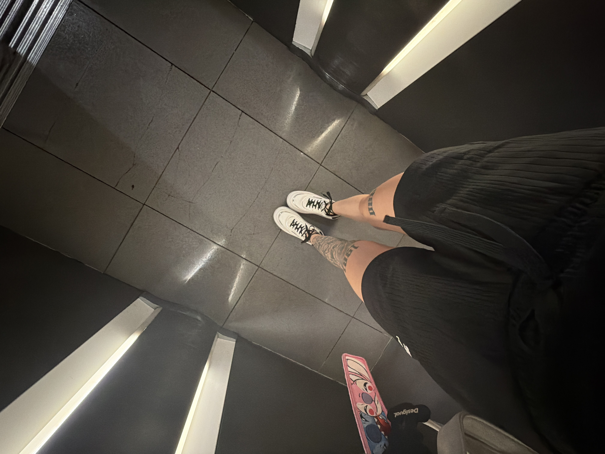 The image, captured from a high-angle overhead perspective, depicts the lower body of an individual standing on a dark grey tiled floor within a modern indoor environment. The person is wearing black, textured shorts and white sneakers with black laces. Tattoos are visible on both shins/calves; the left leg (viewer's right) displays text or numbers, including what appears to be "1983". Adjacent to the individual's left side is a partial view of a light grey stroller or pram, from which a pink accessory featuring Disney's Stitch and Angel characters and the "Desigual" brand logo is visible. The floor is composed of large, rectangular dark grey tiles with lighter grout lines, exhibiting minor wear and reflections from overhead lighting. On both sides of the pathway are dark walls featuring multiple parallel, horizontal white light strips, creating a contemporary and well-lit architectural setting. The scene is located in a modern indoor public space, likely a shopping mall, in Plovdiv, Bulgaria.