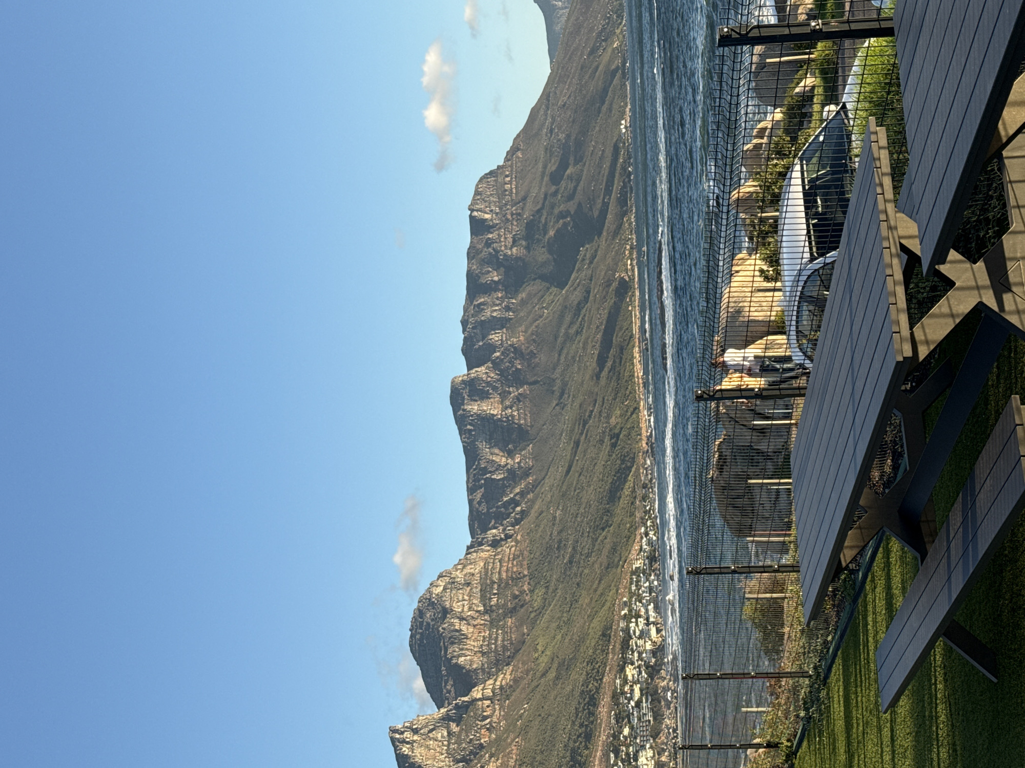 This image features a scenic view of the city of Cape Town, South Africa. A large mountain range dominates the left side of the frame, with clear blue sky. The right side of the frame shows the coastline and the ocean. In the foreground, a black picnic table sits on a patch of green artificial turf, behind a wire fence. A glimpse of a white car is visible through the fence. A city is visible between the mountain range and the ocean.