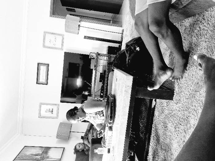 An interior scene captured in black and white presents a family at home. A woman with braided hair leans over a table, focused on an object in front of her. To the side, a young girl reclines on a sofa, possibly engaged with a handheld device. Legs are visible, suggesting someone is nearby, perhaps seated on a high surface. Framed artwork hangs on the wall, adding a personal touch to the space. The carpet adds texture and depth.