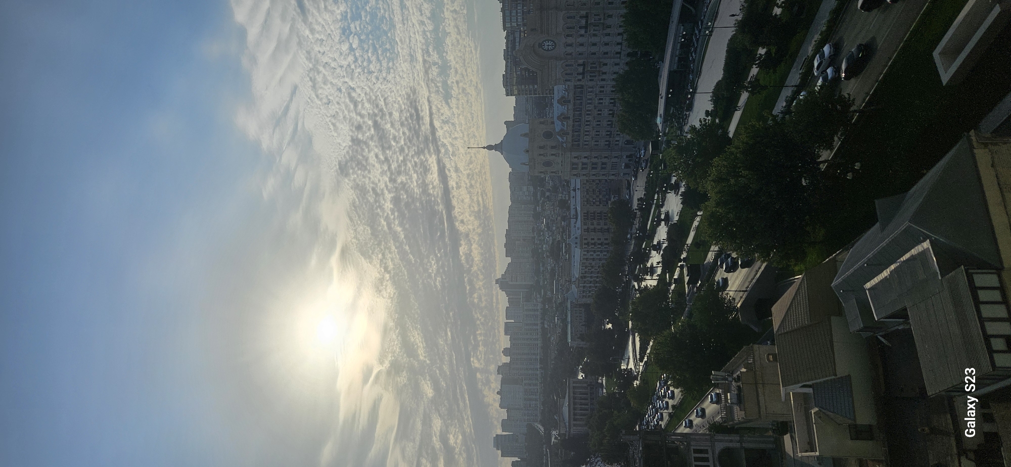 This is an aerial, vertical shot of the city of Baku, Azerbaijan, during daytime, likely early morning or late afternoon, given the low sun angle and hazy light. The scene overlooks a wide boulevard lined with trees, grassy areas, and several grand buildings, one with a prominent dome and clock tower. Traffic is visible on the boulevard, with cars moving in both directions. In the background, a dense cityscape of apartment buildings stretches across the horizon under a sky filled with wispy, patterned clouds. The foreground shows rooftops of residential buildings. The image is watermarked "Galaxy S23".