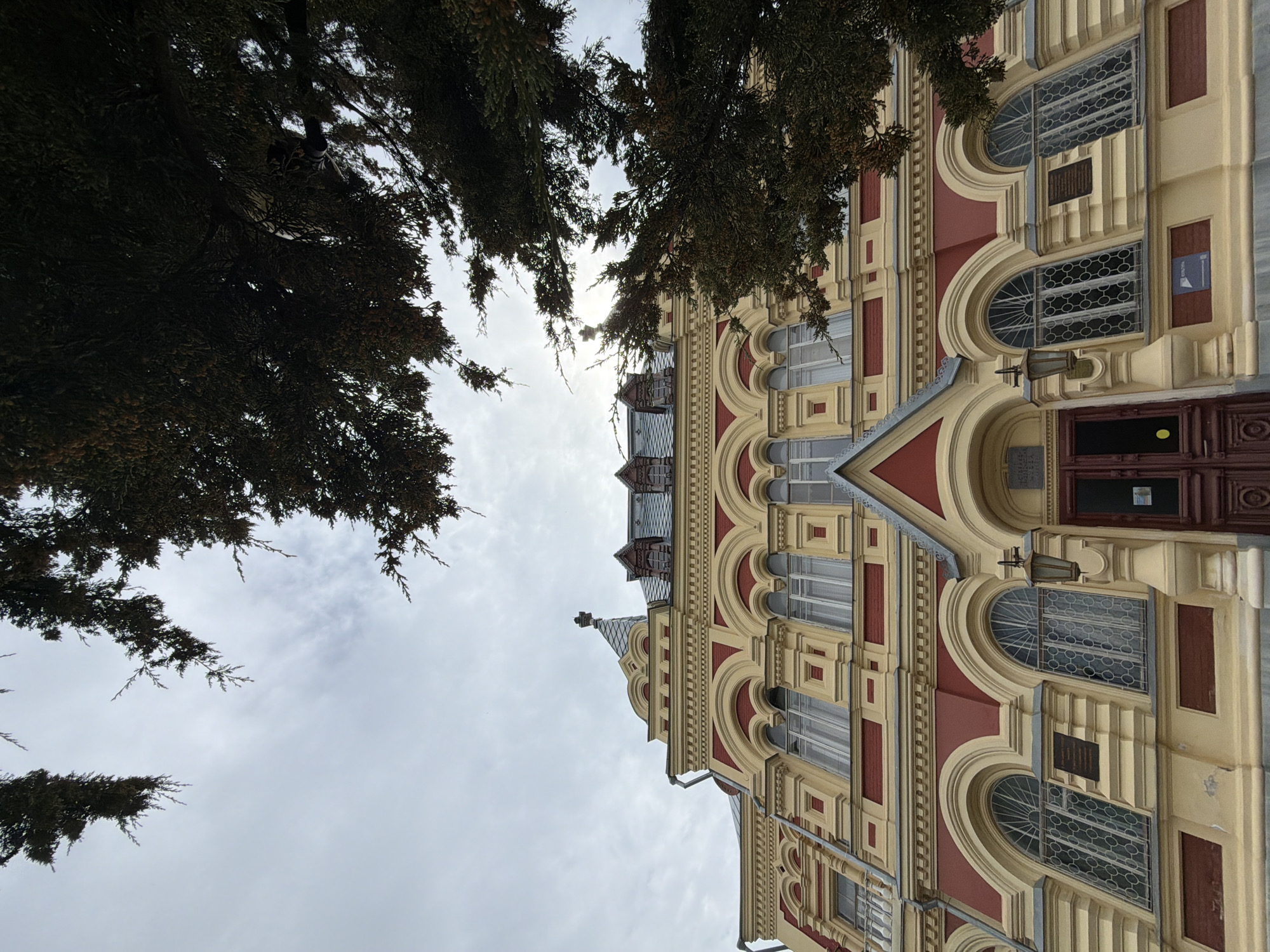 The image depicts the Kamyshin Historical and Local Lore Museum in Kamyshin, Russia. The view is from a low angle, looking up at the facade of the building, which is partially obscured by tree branches on the left side. The sky is overcast. The building is a multi-story structure with a distinctive architectural style, featuring arched windows, ornate decorative elements in red and yellow colors. The main entrance is visible with a brown door. Various plaques and small decorative lights are mounted on the facade. The top of the building has a roof with metal accents.