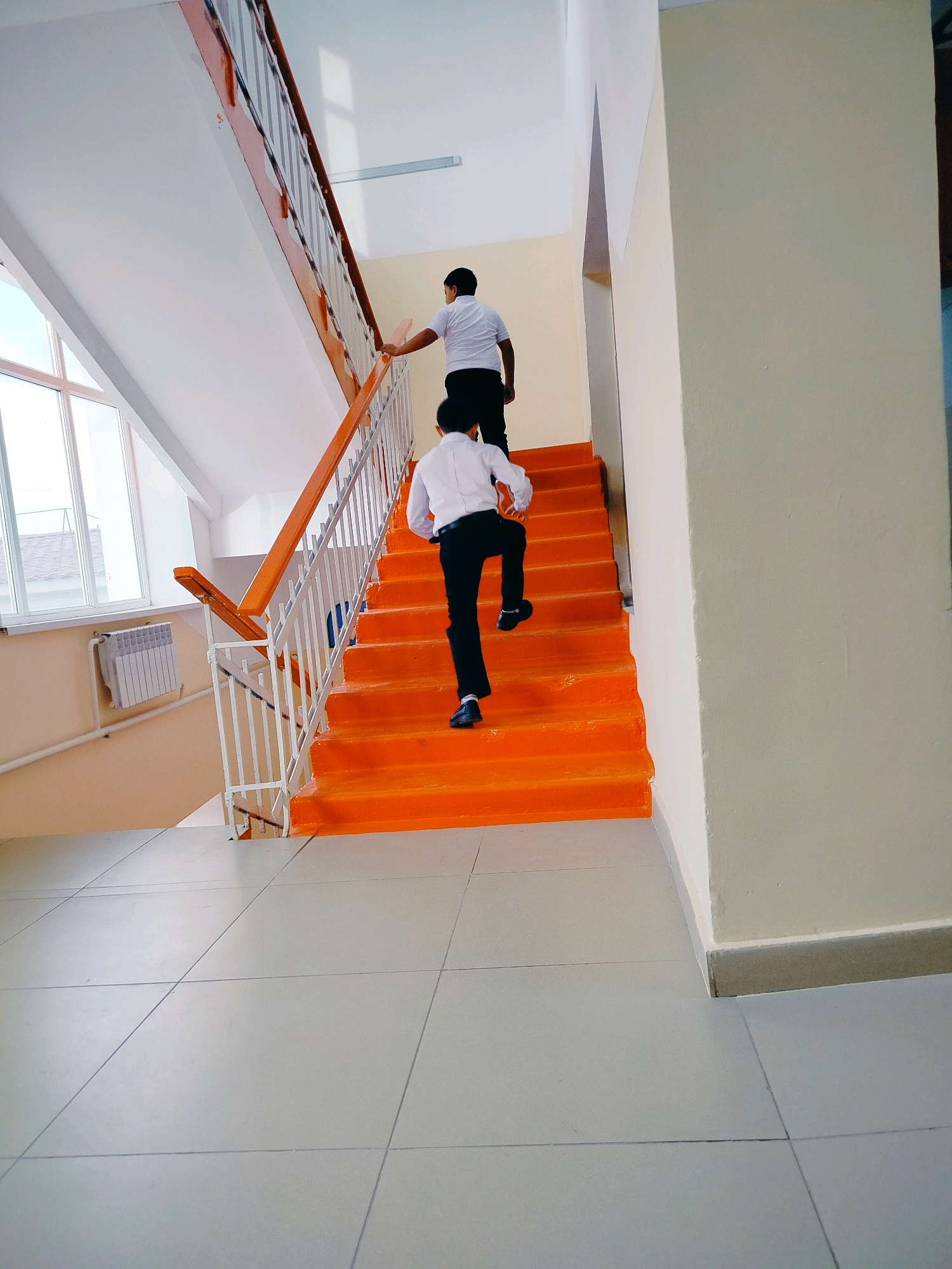 Two young males, appearing to be school-aged boys, are ascending a bright orange staircase. The boy in front is wearing a white collared shirt and black pants, with dark shoes, and is in mid-stride, his right leg extended. The boy behind him is also wearing a white collared shirt and black pants, and is further down the stairs, with his right leg bent as if he is about to take a step. The staircase has white metal railings with a wooden top handrail, painted orange along the sides. The walls are painted in a light cream color, and the floor is tiled with large, light grey square tiles. To the left, a large window allows natural light into the stairwell, and a white radiator is mounted on the wall below it. The overall environment suggests an interior space, possibly a school or public building. The image location is identified as Kölkent, Kazakhstan.