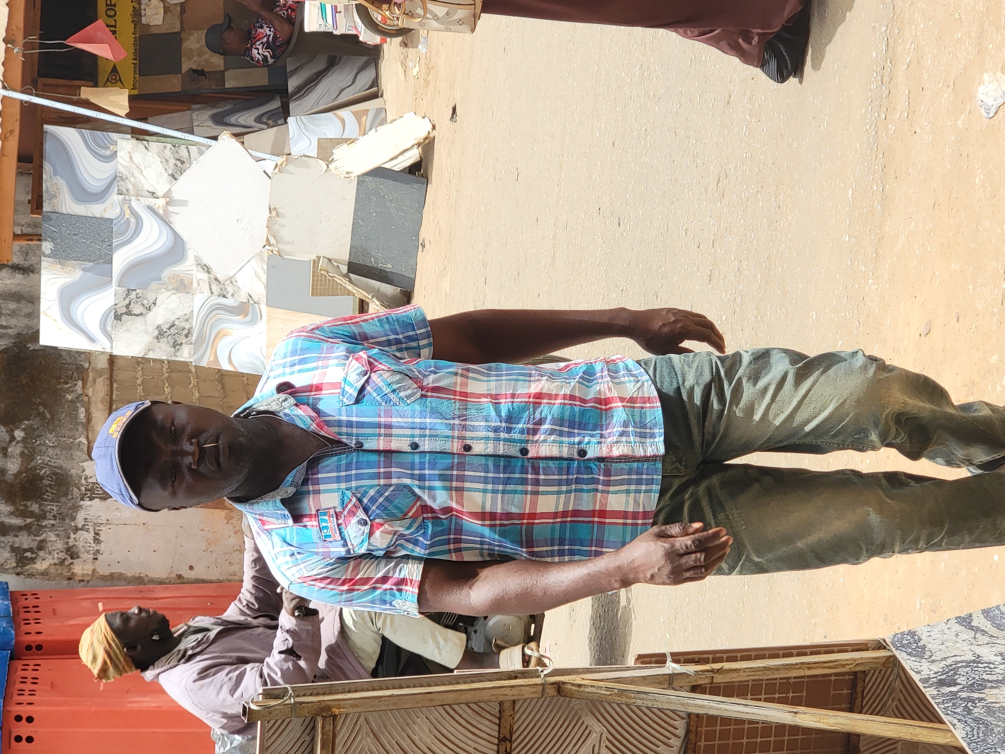 A street scene in Maiburiji, Nigeria, shows two adult Black men standing in front of a stack of construction tiles. One man wears a blue baseball cap, a blue and red plaid shirt, and dark jeans. A toothpick is in his mouth. The second man, wearing a head wrap, is slightly behind and to the left. In the background are various tiles stacked against a wall and displayed in front of a shop. The ground is a tan, sandy color, indicating an outdoor environment.