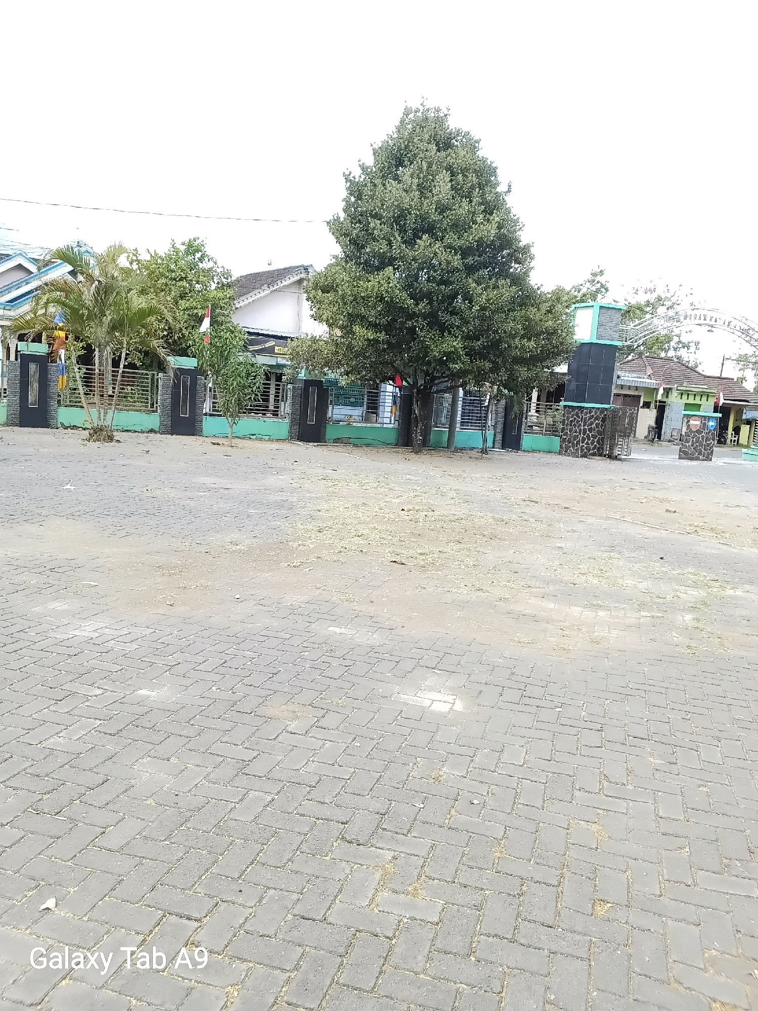 The image shows a paved courtyard in front of a building complex, possibly a school or community center, in Karangan, Indonesia. The courtyard is paved with interlocking grey bricks laid in a herringbone pattern. Scattered on the paving are small amounts of dry grass and debris. In the background, a fence with vertical black posts and horizontal metal bars separates the courtyard from the buildings. The buildings have green walls and grey tiled roofs. A large, leafy tree stands prominently in the middle of the background, with other smaller trees and palm trees visible. To the right, a large, multi-faceted speaker system is mounted on a dark structure. An archway with white lettering is visible further in the background, above the speaker. Two Indonesian flags are visible, one red and white flag on a flagpole and another red and white flag attached to a building. The sky is overcast and bright white. The text "Galaxy Tab A9" is visible in the bottom left corner of the image.