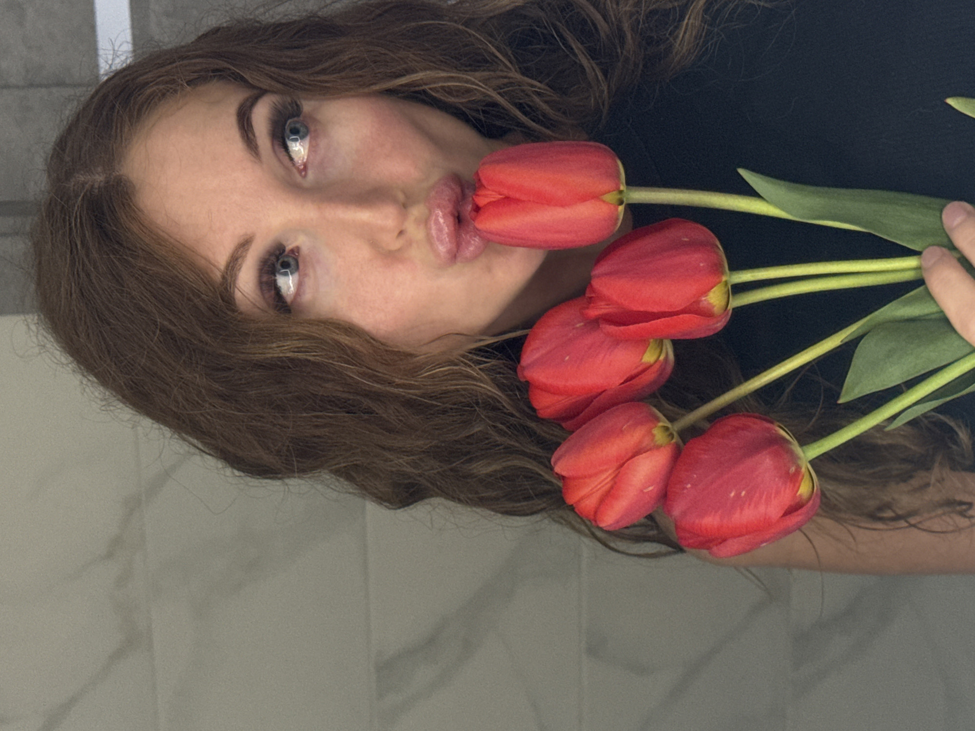 A woman with light skin, blue eyes, and brown hair holds a bouquet of four red tulips in her right hand and poses with one tulip between her lips. She wears a black top. The background consists of a wall with a light grey marble tile pattern. It is not possible to determine from this image if this photograph was taken in the city of Chaikovsky, Russia.
