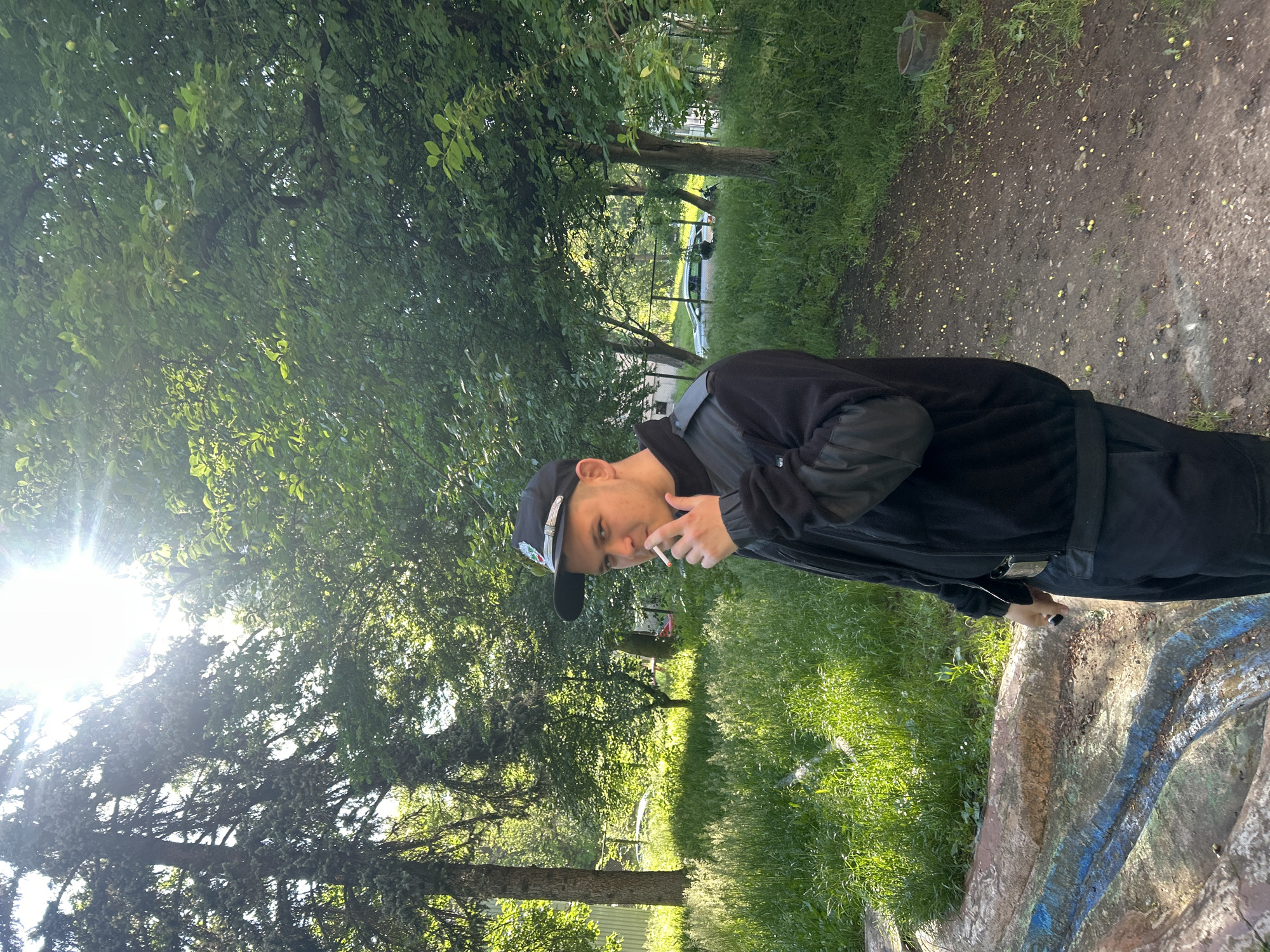 A medium, eye-level shot, rotated sideways, features a man standing outdoors in a park-like setting. The man is wearing a black cap, a black jacket, and dark-colored pants. He is holding a lit cigarette between his fingers near his mouth. The background consists of a variety of trees, grassy areas, and a pathway. Sunlight filters through the foliage, creating areas of light and shadow. A painted concrete barrier or wall is visible in the foreground.