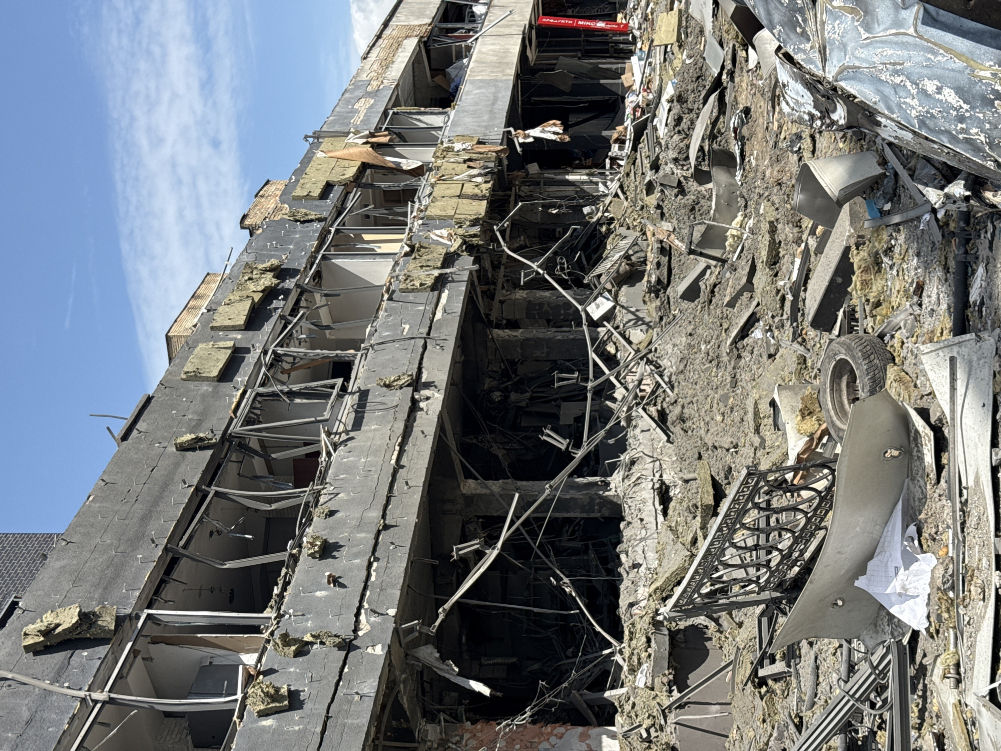 The image shows a severely damaged building in Kyiv, Ukraine, post-attack. The building's facade is heavily scarred with large sections missing or crumbling. Window frames are twisted and shattered. Debris is scattered around the base of the building, including twisted metal, insulation material, and structural elements of the building. A car component and decorative railing are visible amid the rubble. The sky above is clear and blue. The building's sign partially showing the words "ПРОДУКТИ МИКО..." can be seen at the top.