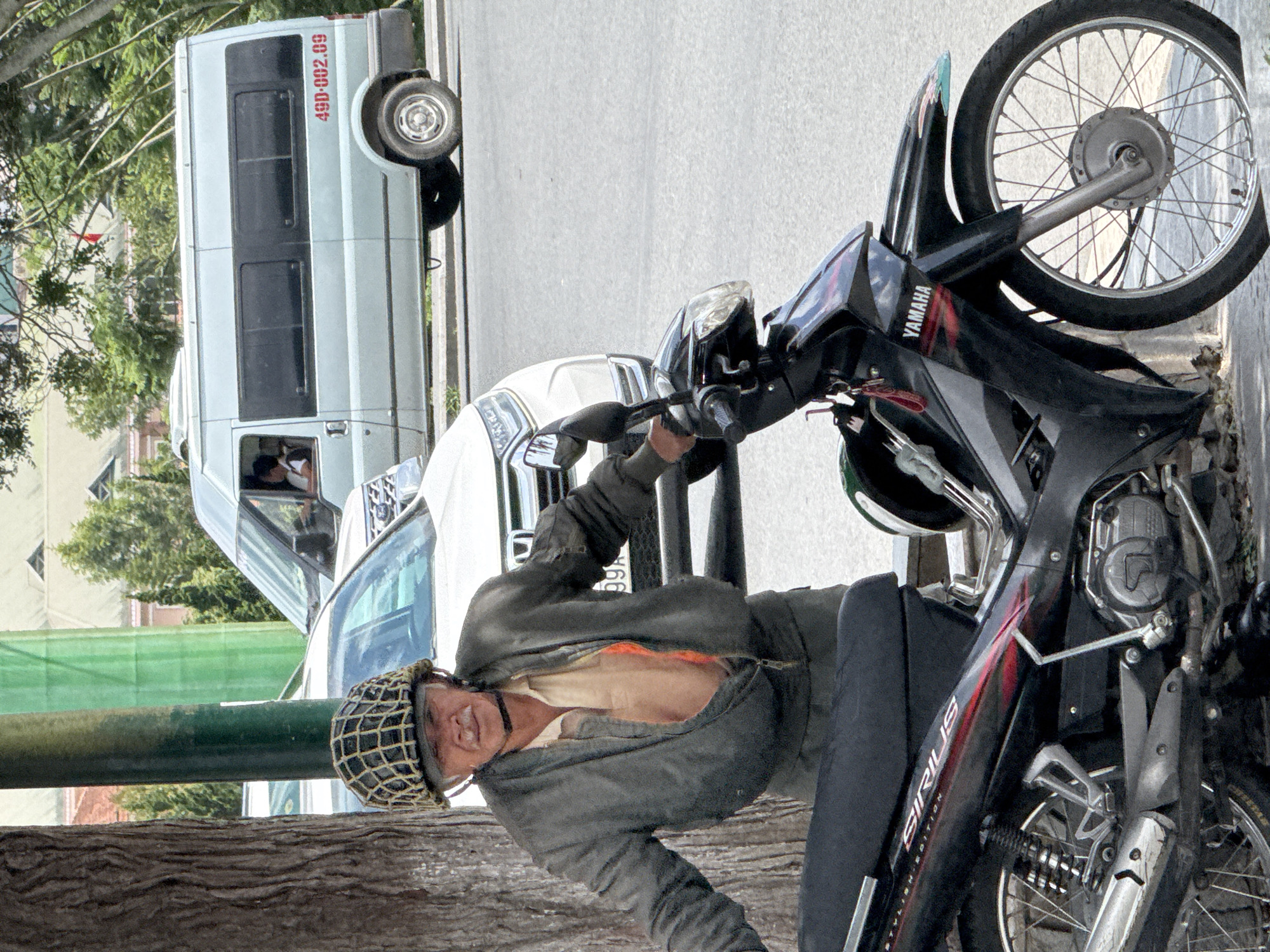 In Da Lat, Vietnam, an elderly Vietnamese man, wearing a distinctive netted helmet and a gray bomber jacket unzipped to reveal a tan collared shirt with an orange undershirt, leans on his black Yamaha Sirius scooter. The scooter has red and black accents on its body, and a key with a red tag hangs from the handlebars. Behind the man and his scooter, a light blue passenger van with the license plate "49D-002.09" is parked on the street. A white SUV is positioned between the man and the van, partially obscured. The background reveals lush green trees and a building with a light-colored roof. The scene appears to be a roadside in Da Lat, characterized by a mix of vehicles and the natural environment.