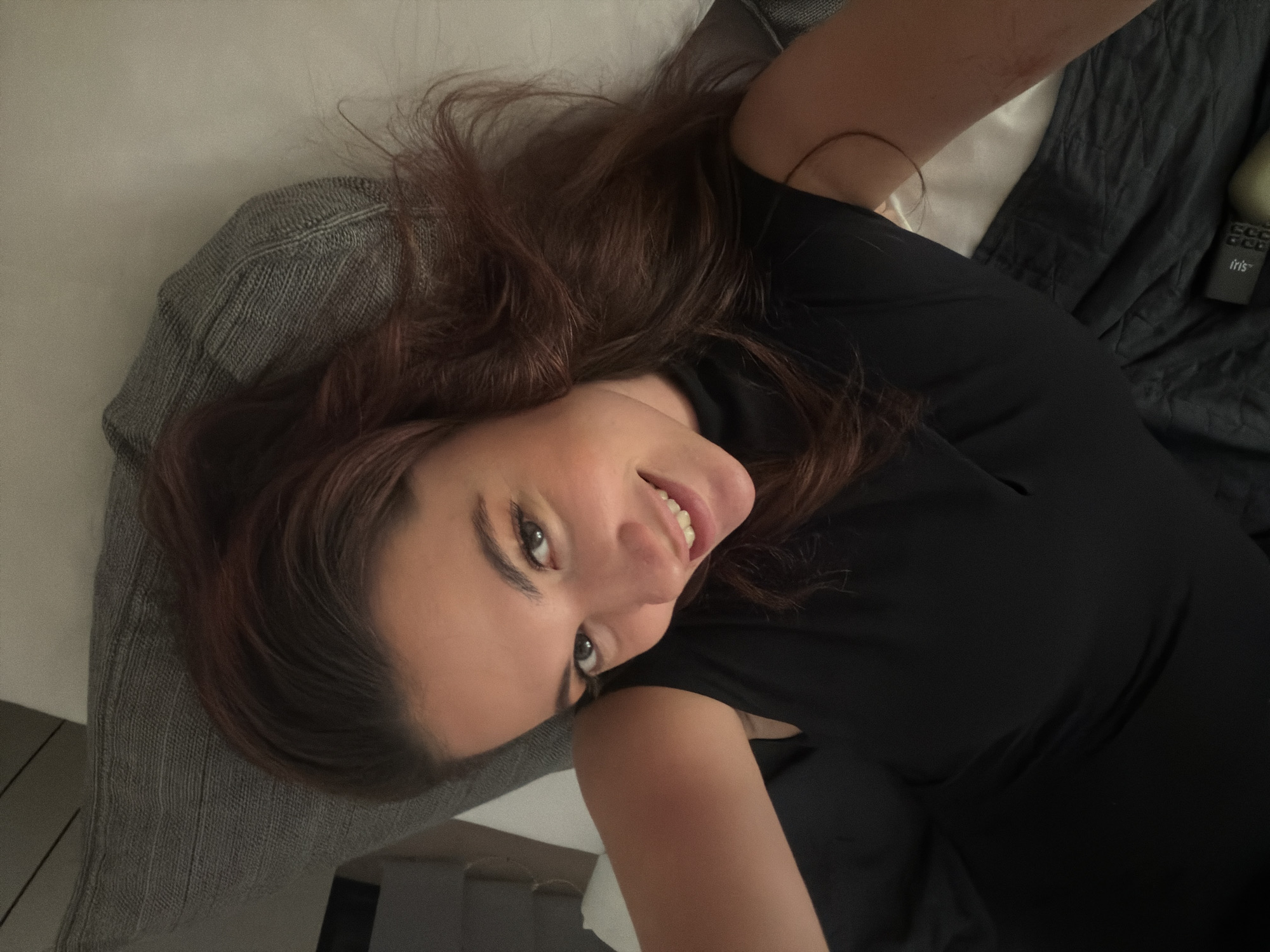 The scene depicts a female individual in a close-up, high-angle view, situated indoors, likely within a private residence or hotel room in Belgrade, Serbia. The subject is lying or reclining and is looking directly at the camera with a smile, teeth visible. She has dark brown hair and is wearing a dark, sleeveless top or dress with a high neckline. The background includes a textured grey pillow located to her upper left, a light-colored sheet or surface to her right, and a dark, textured fabric, consistent with a blanket or bedspread, covering the lower right portion of the frame. A remote control, partially visible on the dark fabric, features the brand name 'Iris' and several control buttons. Adjacent to the remote, a light-colored, cylindrical object is also partially visible.