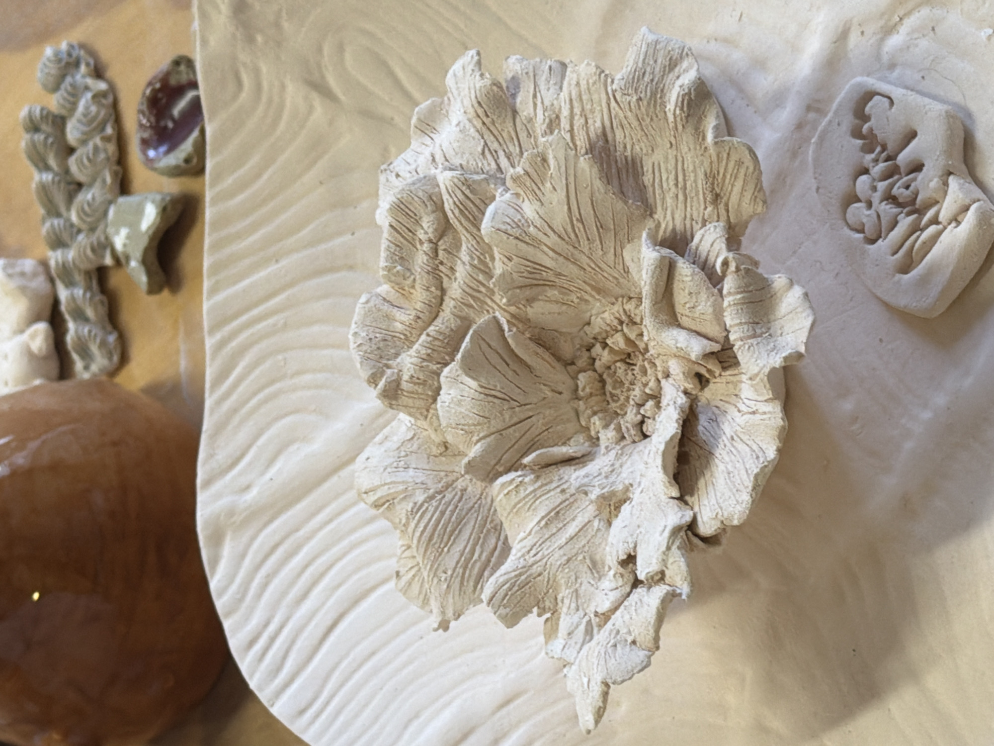 The image displays an arrangement of ceramic art pieces, primarily featuring a large, textured flower relief at its center. This central piece is off-white and depicts a blooming flower, possibly a peony or camellia, with numerous overlapping petals detailed with fine, parallel lines that give a sense of organic structure and texture. The center of the flower is heavily textured and intricate.
The flower relief is set against a flat, light-colored background surface. To the left and below the main flower, this background has a distinct wavy or ridged pattern, possibly created by tool marks or a texture press. To the right and above, the background appears smoother but still has a slightly uneven, matte finish.
Various other ceramic elements are scattered around the main flower. On the far left, partially visible, is a cluster of smaller, gray-toned ceramic pieces, one resembling a series of braided or coiled shapes, and another an irregular fragment. Above these, a small, dark reddish-brown, polished object, possibly a stone or glazed ceramic piece, is present. On the upper right, a distinct off-white ceramic mold or cast features an intricate, organic pattern within its border, possibly representing seeds or small florets. A large, shiny, reddish-brown, rounded object is partially visible on the far lower left.
The overall impression is a collection of handcrafted ceramic works, possibly showcasing different stages of the artistic process or a variety of textural studies. The lighting is soft and even, highlighting the textures and three-dimensionality of the pieces. There are no people visible, nor any discernible text.