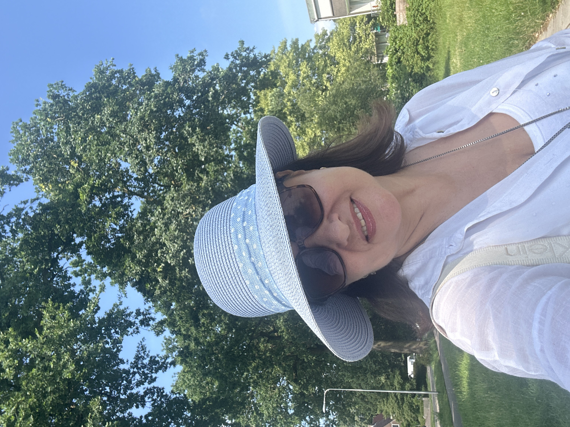 A woman is taking a selfie outdoors. She is wearing a blue straw hat with a white polka dot ribbon, sunglasses, and a white linen shirt over a white tank top. A silver necklace is visible. She is standing in front of large green trees and a bright blue sky. In the background, there is a street with a lamppost, houses, and green grass. The image appears to be taken in a suburban or park-like setting in Steenwijk, Netherlands.