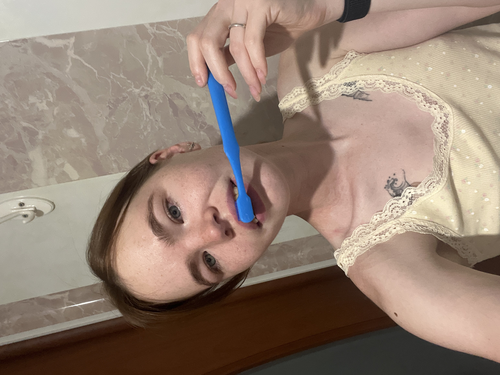 A young Caucasian woman with light skin, freckles, and light brown hair is brushing her teeth in a bathroom. She is holding a blue toothbrush in her right hand and has it in her mouth. She is wearing a light yellow tank top with lace trim and a floral pattern. There are two tattoos visible on her chest. The background appears to be a bathroom with marble-patterned tiles and a light-colored wall with a hook. The image is taken from a medium-close-up perspective, focusing on the woman's face and upper body. The lighting suggests an indoor setting, possibly a bathroom at night. The location is Chaikovsky, Russia.