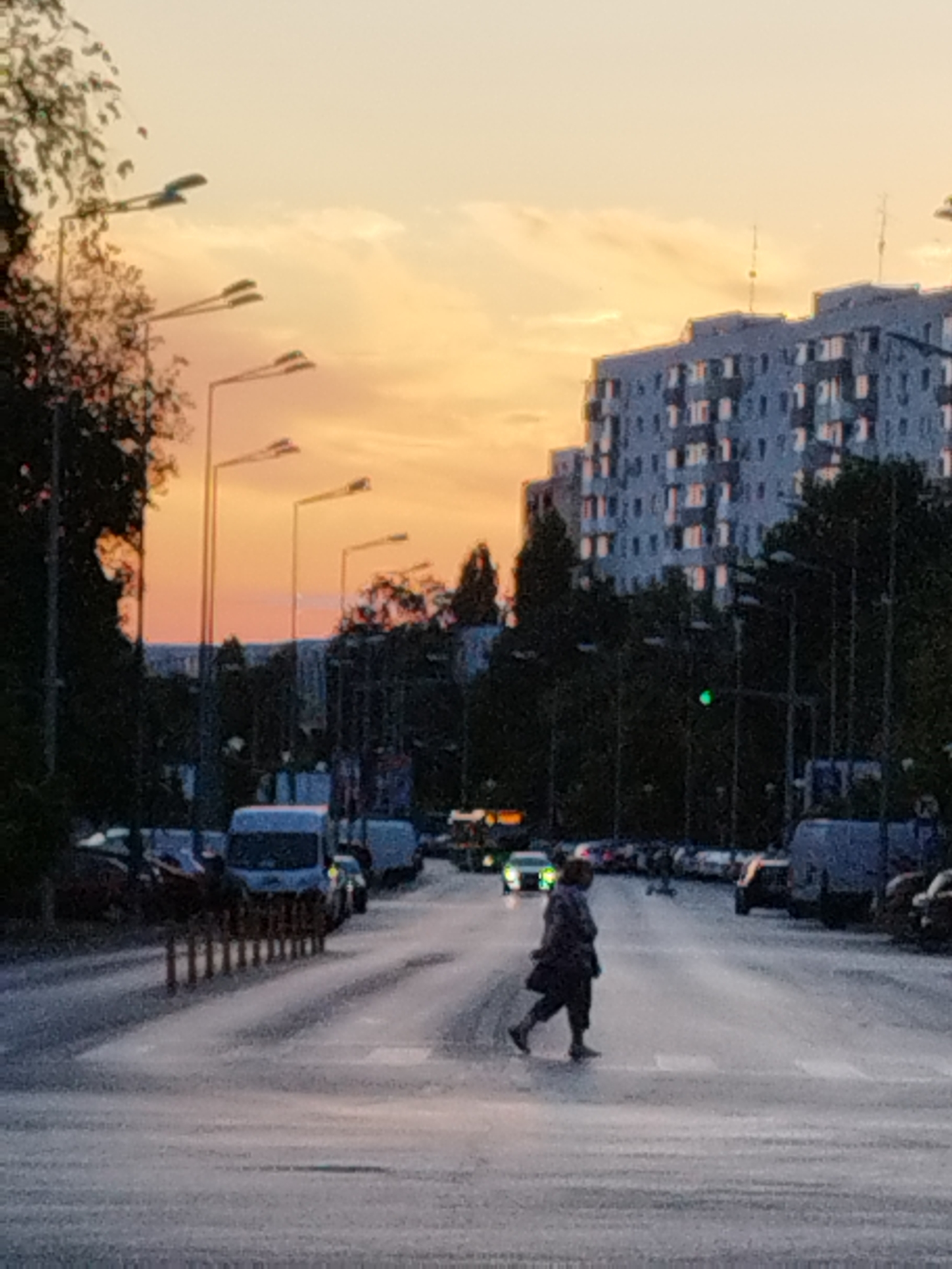The image depicts a street scene in Bucharest, Romania, during twilight. A pedestrian, dressed in dark clothing, is crossing a street at a crosswalk. The sky is a gradient of orange and yellow, suggesting a sunset or sunrise. In the background, a multi-story apartment building with a distinctive tiered architecture dominates the skyline. Streetlights are visible along the side of the road. Several vehicles, including vans and cars, are parked or driving on the street. The scene appears to be in a residential or mixed-use area of the city.