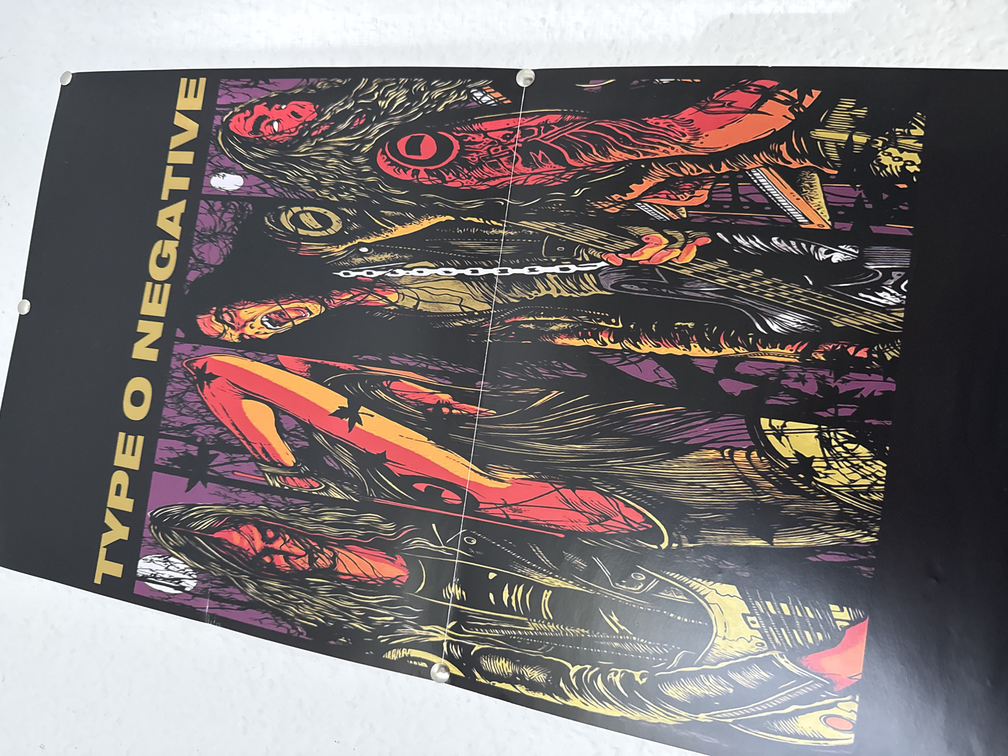 The image is a photograph of a poster for the band Type O Negative. The poster features an illustration of the band members in a dark, forest-like setting. The band members are depicted with long, disheveled hair and intense expressions. They are dressed in dark, stylized clothing, with chains and other accessories. The color palette of the poster is dominated by black, red, yellow, and purple, creating a gothic and macabre aesthetic. The poster is hung on a white wall, and there are metal fasteners visible at the top corners and along the sides, suggesting it is pinned or mounted. The lighting in the photograph is even, with no strong shadows. The image is cropped to focus on the poster itself. There is no visible indication of any specific location in Bad Sachsa, Germany.