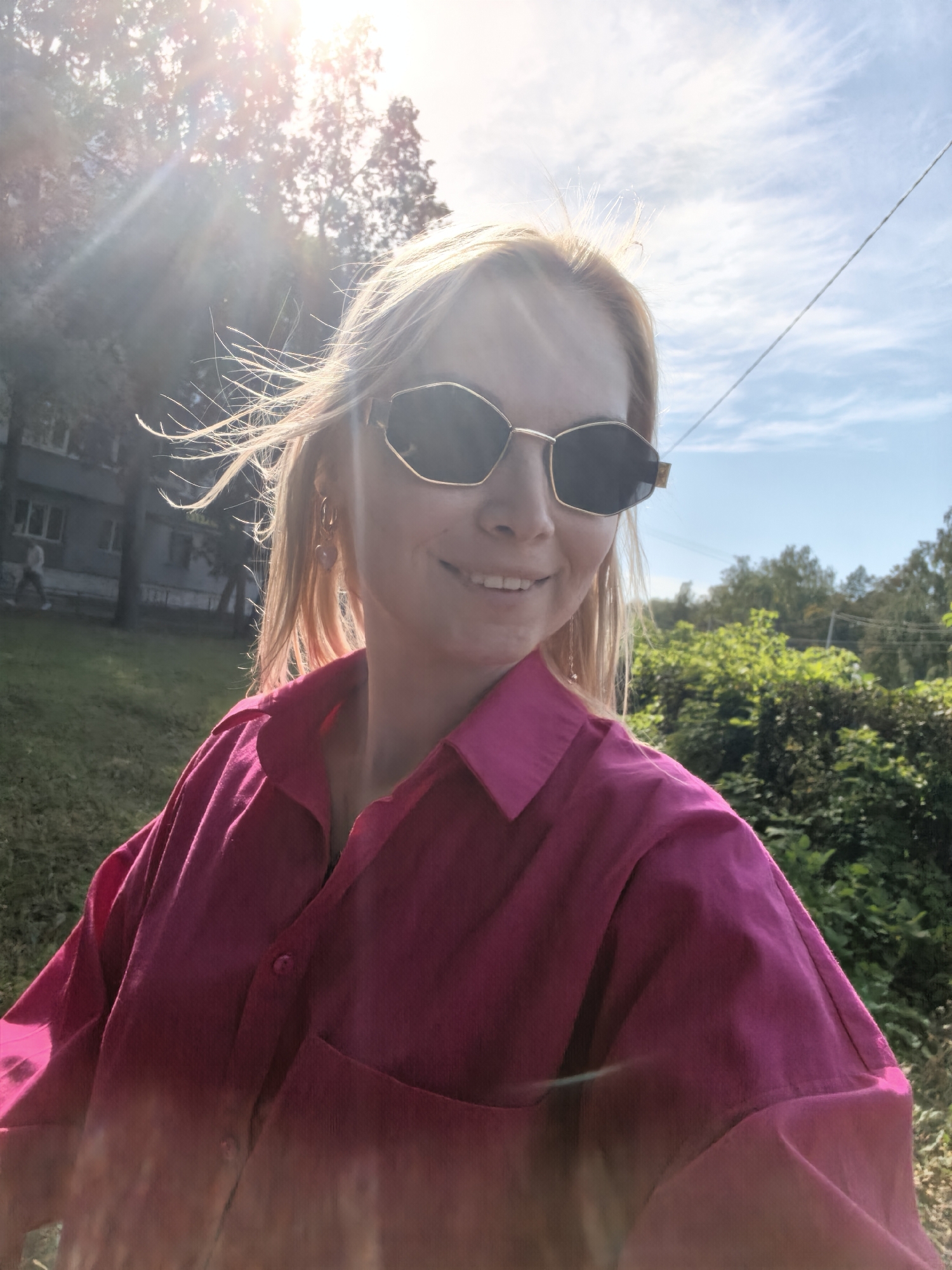 A young woman with blonde hair and sunglasses is pictured from the chest up, outdoors in what appears to be a park or residential area. She is wearing a bright pink, button-down shirt. Her hair is blowing in the wind. The sun is shining brightly, creating lens flare in the upper left corner of the image. In the background, there is a building, trees, and a blue sky with scattered clouds. The image appears to have been taken on a sunny day.