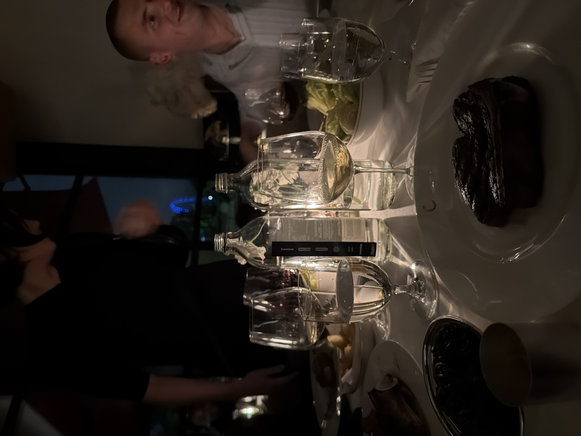 The image depicts a dimly lit interior scene of a restaurant, likely in London, United Kingdom, given the presence of the London Eye visible in the background through a window. The table is set for a meal, with white plates, wine glasses, and bottles of water. A steak is prominently featured on the plate in the foreground. In the background, several individuals are visible, including a person in a black shirt and another man smiling. The atmosphere is intimate, with soft lighting that casts shadows and highlights the objects on the table. The overall impression is one of a fine dining experience.