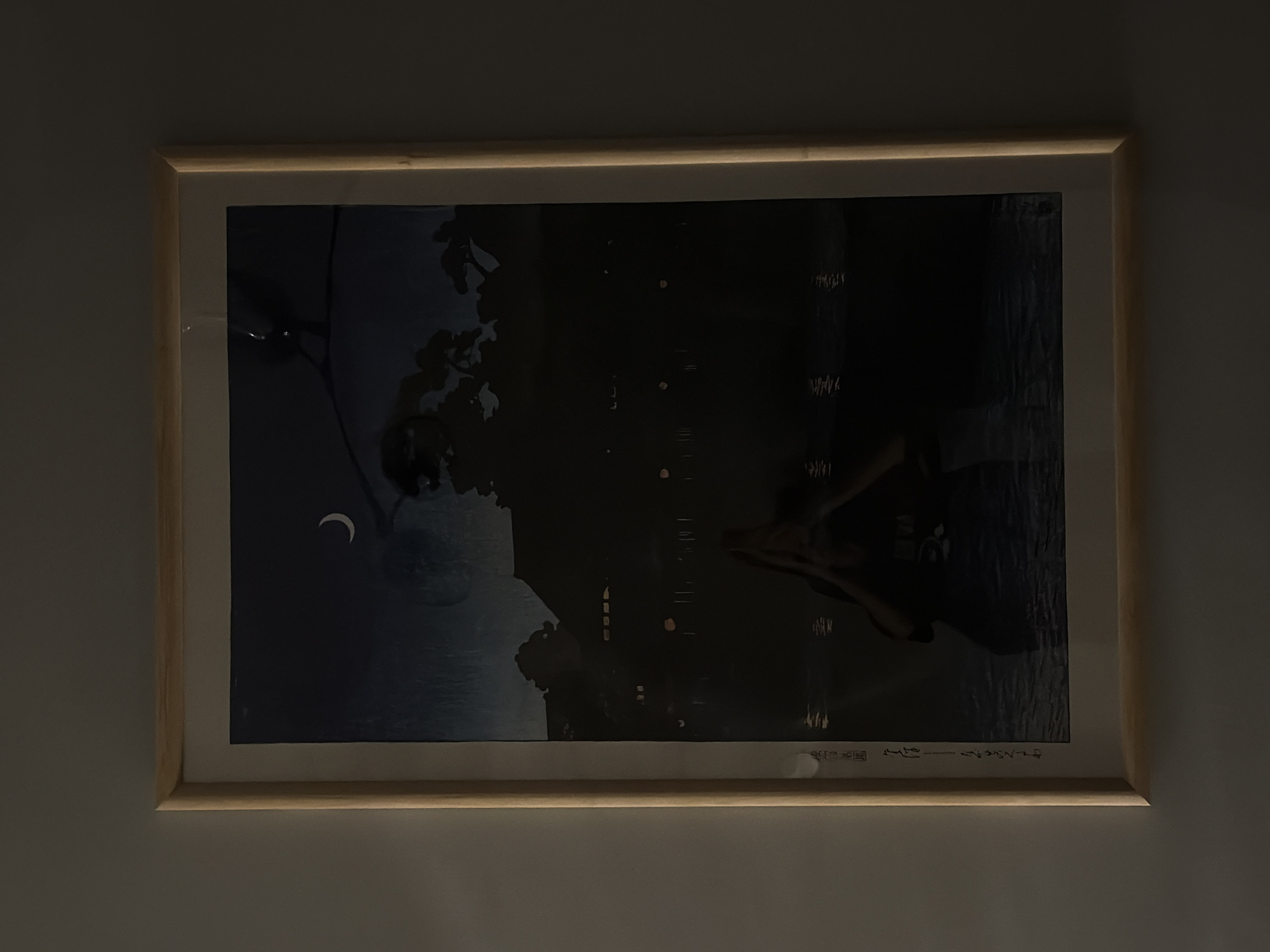 A framed artwork hangs on a light gray wall. The artwork depicts a dark scene with a crescent moon in the upper left. There are tree branches above the moon, and below there appears to be water reflecting lights from the city. The title and the artist's name are written in Japanese along the bottom of the artwork. The frame appears to be light-colored wood. The location in the city of Khimki, Russia cannot be determined from the image.
