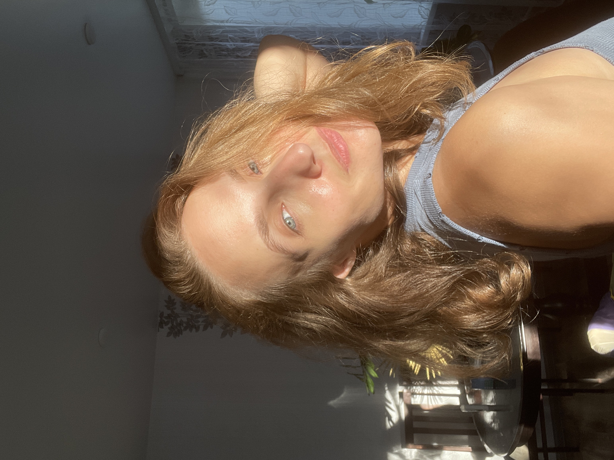 This media file presents a close-up portrait of a woman, rotated 90 degrees counter-clockwise. The woman, appearing to be in her 20s or 30s, is the central subject, looking directly into the camera with a calm expression. She has light brown to blonde, wavy hair that frames her face and shoulders. Her eyes are light blue or green, and her lips have a natural, slightly pink tone. Her skin appears fair and radiant, with highlights suggesting a natural sheen. She is wearing a light blue or grey ribbed tank top, with her shoulder and upper chest visible.
The scene is an indoor setting, likely a room in a residence in Sharkan, Russia. Strong, direct sunlight streams in from what appears to be a window or blinds located in the upper right portion of the frame (as oriented). This light creates bright highlights on the woman's hair, forehead, nose, and shoulders, contrasting sharply with deep shadows in the background and on other parts of her body. Shadows of window frames or blinds are visible on the wall and ceiling above her head.
The background consists of a plain wall, mostly in shadow in the upper left, where a faint circular object (possibly a light fixture or wall decor) can be discerned. In the lower left, shadows resembling leaves or a plant are cast onto the wall. The lower right shows indistinct, shadowed furniture, possibly part of a table and chairs, with some reflective surfaces catching the light.
The strong, directional lighting indicates it is daytime, likely morning or late afternoon. The overall impression is one of natural light and a relaxed atmosphere. There is no visible text in the image.