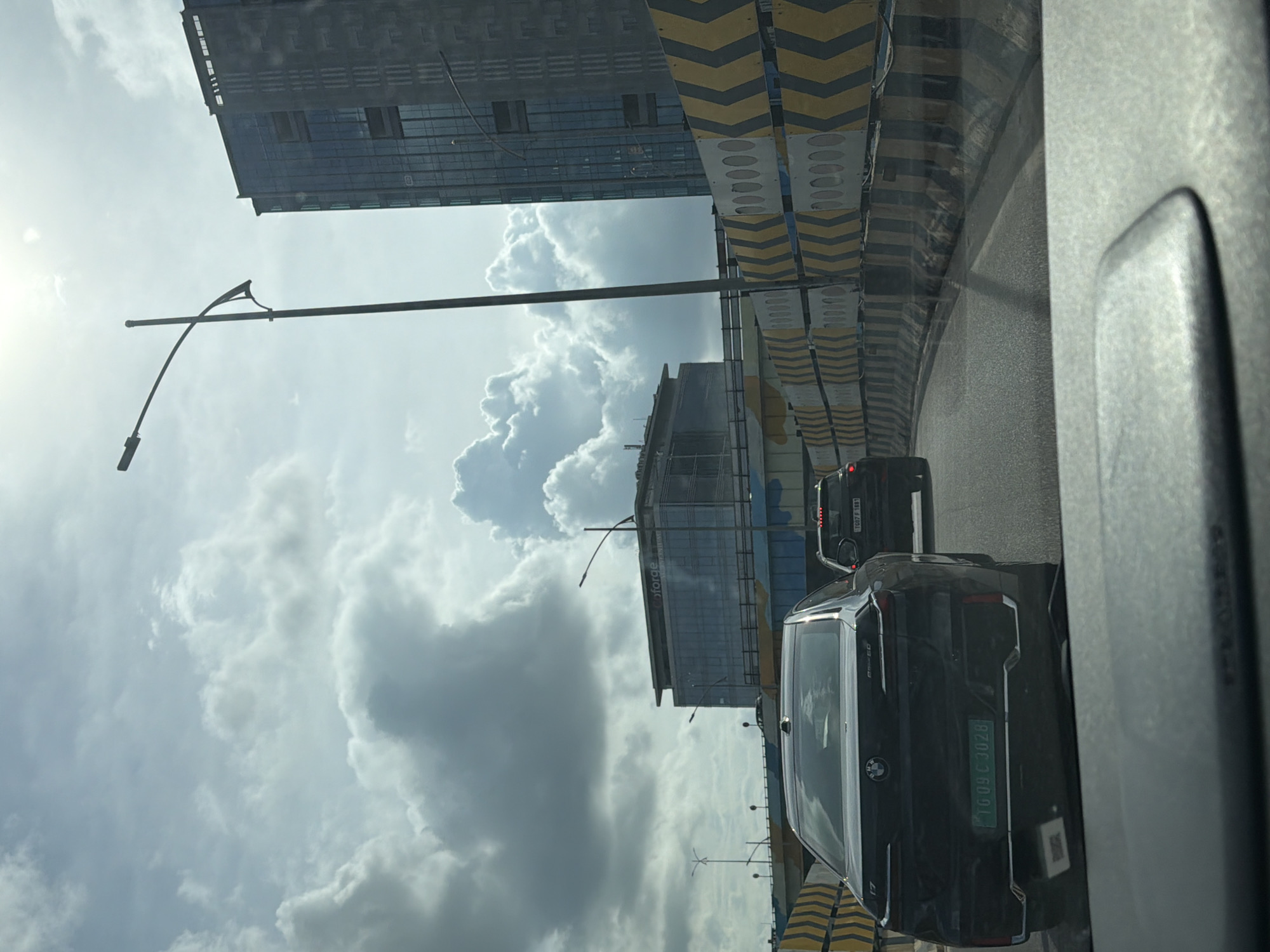 In Hyderabad, India, a black BMW car is viewed from the interior of another vehicle, driving on a highway. The BMW has a green license plate that reads "TG 09 C 3028". Behind it, another car is visible, its license plate partially obscured but showing "TG 07 F 1881". The highway is flanked by concrete barriers with alternating yellow and black chevron patterns. In the background, two modern buildings are under construction or recently completed. The larger, taller building on the right is a glass-fronted skyscraper with visible vertical architectural elements. To its left, a lower, wider building with "Infosys" branding on its facade is also visible. The sky is partly cloudy with bright sunlight.