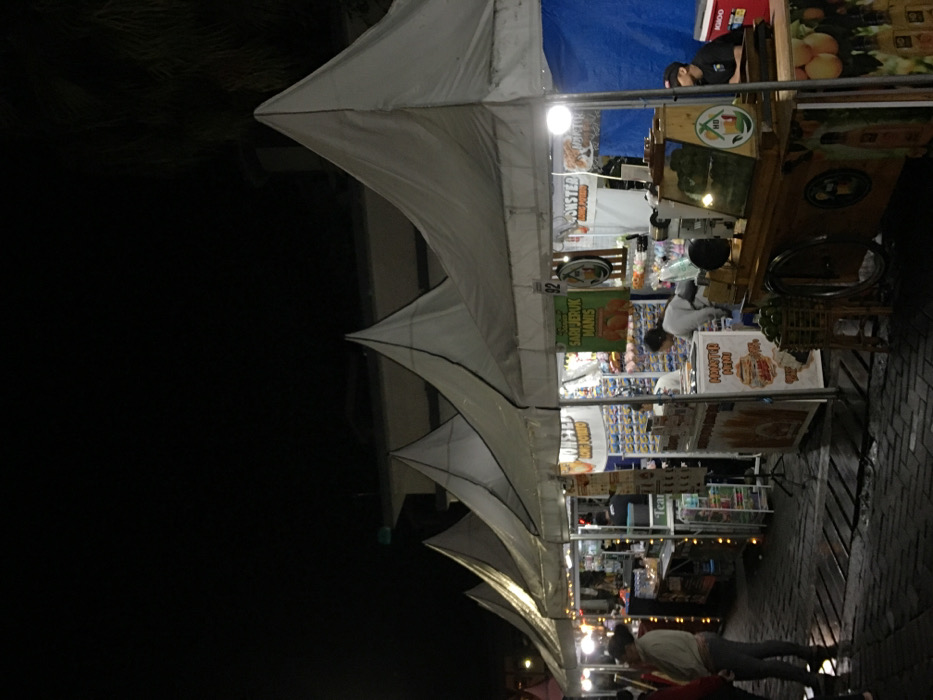 A nighttime view shows a street market lined with vendor stalls under white peaked canopies. People browse and work at the stalls, which are stocked with various goods and food items. Signage is visible, advertising "Sari Jeruk Manis" (sweet orange juice) and other products. The scene is illuminated by overhead lights, and the ground is paved with bricks. The overall atmosphere suggests a vibrant and busy outdoor market.