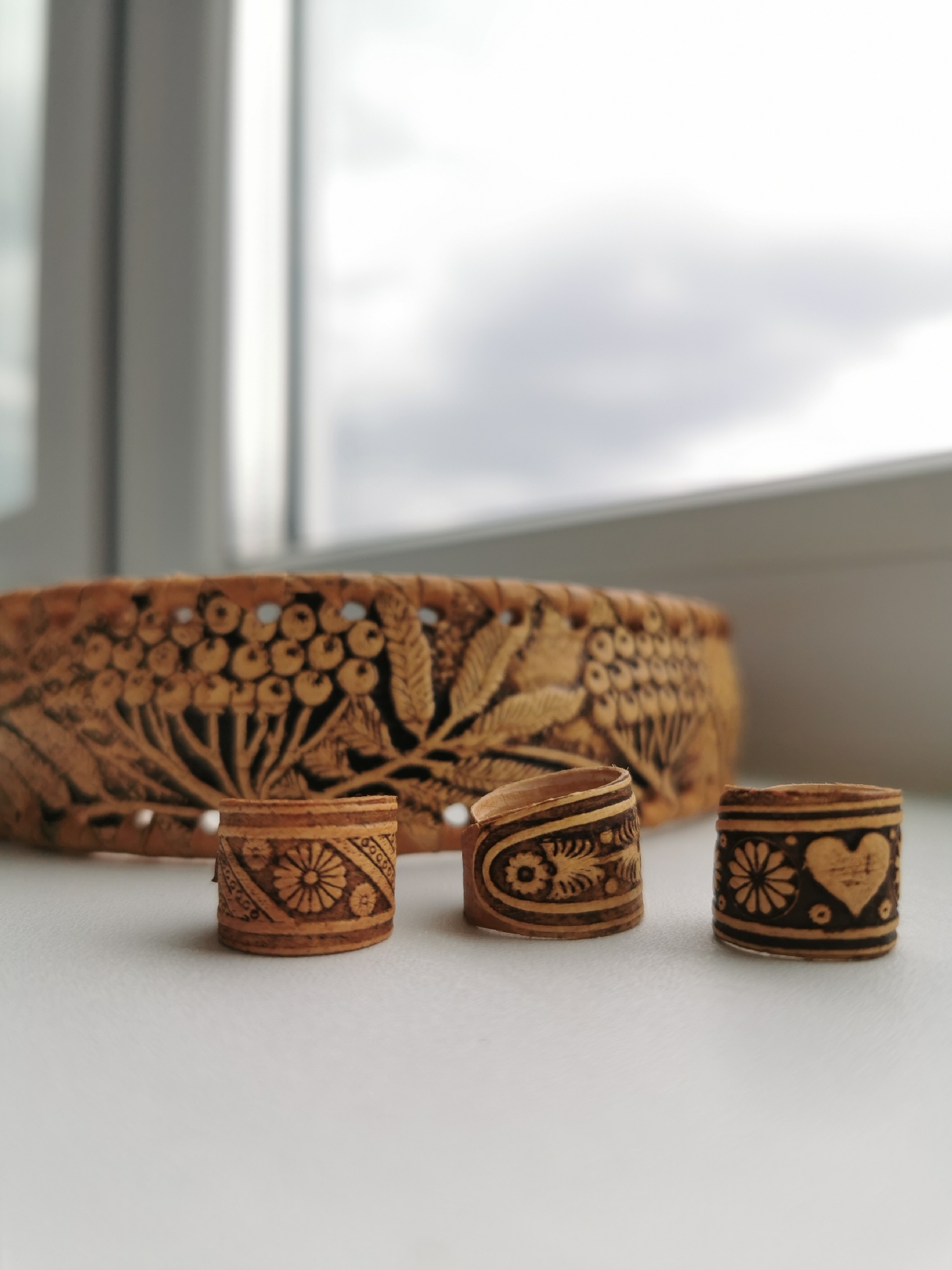 The image captures a still life arrangement of handcrafted items, likely made from birch bark or a similar light wood. In the foreground, three small, cylindrical objects are neatly aligned on a light-colored, possibly white, surface. These appear to be decorative rings or thimbles, each featuring unique, intricate dark carvings against the natural wood tone.
The leftmost small cylinder displays a central stylized flower surrounded by geometric patterns and small circles. The middle piece has a more fluid, organic design with swirling leaf or vine motifs, and it appears slightly open or curved. The rightmost cylinder features a prominent heart shape and a floral design, also accented with small circles and lines.
Behind these small items, a larger, shallow basket or bowl-like object is visible, partially in focus. This larger piece is elaborately carved with a continuous pattern of what appears to be clusters of berries (resembling rowan berries) and leaves, with parts of the design cut out to create a lacy effect. The dark carvings contrast with the light, natural color and texture of the bark material.
The background consists of a blurred window view, indicating an indoor setting. Through the window, an overcast or cloudy sky is visible, suggesting natural, diffused daylight. No specific activity is occurring; the image is a close-up, showcasing the craftsmanship and decorative details of the wooden objects. There is no visible text in the image.