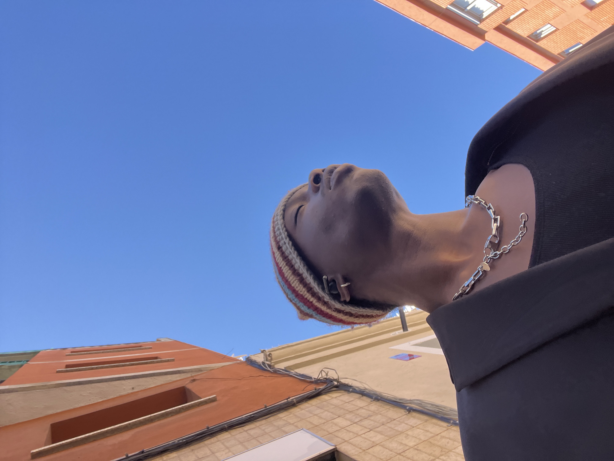 The image shows a low-angle perspective looking up at a person with their head tilted back. The person wears a striped knit cap and a silver chain necklace. The person is wearing black clothing. The background features a bright blue sky and the facades of buildings with orange and beige tones. Based on the architectural style and building colors, it is possible the image was taken in Barcelona, Spain. There is no information about the actions and interactions between the people or objects.