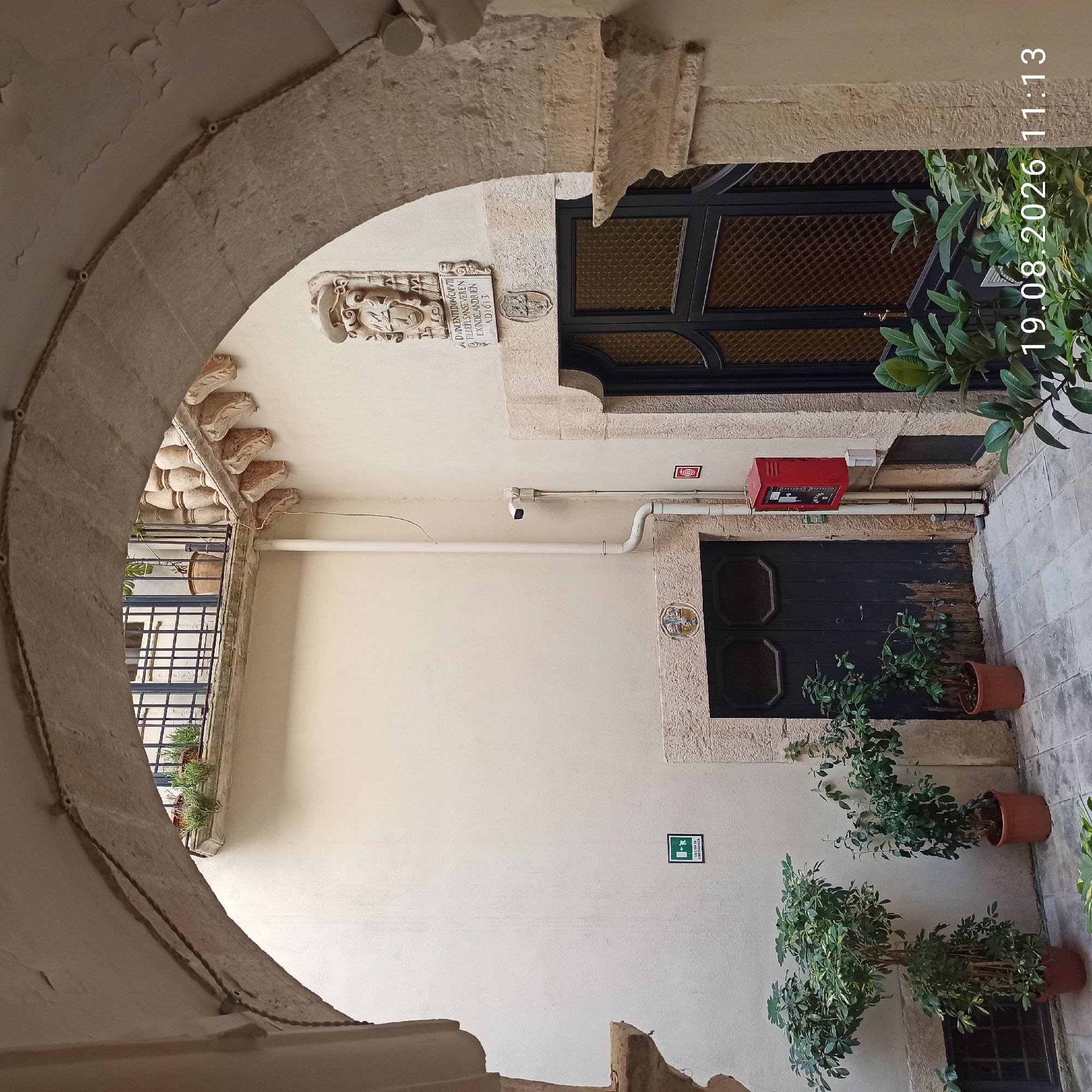 The image captures a view of an inner courtyard in Ruvo di Puglia, Italy, framed by a stone archway. The courtyard features aged plaster walls, stone detailing around doorways, and a tiled floor. On the left, several potted plants are arranged, including a tall Schefflera. To the right, a dark wooden double door is visible, above which is a stone plaque with an embossed crest. Further right, a dark metal door with patterned glass panes stands next to another stone plaque inscribed with text and crests, dating the structure to AD 1613. A red emergency box and a security camera are mounted on the wall. An overhead balcony with a metal railing and potted plants can be seen in the background, above the arch. The overall ambiance suggests a historical setting within the city.