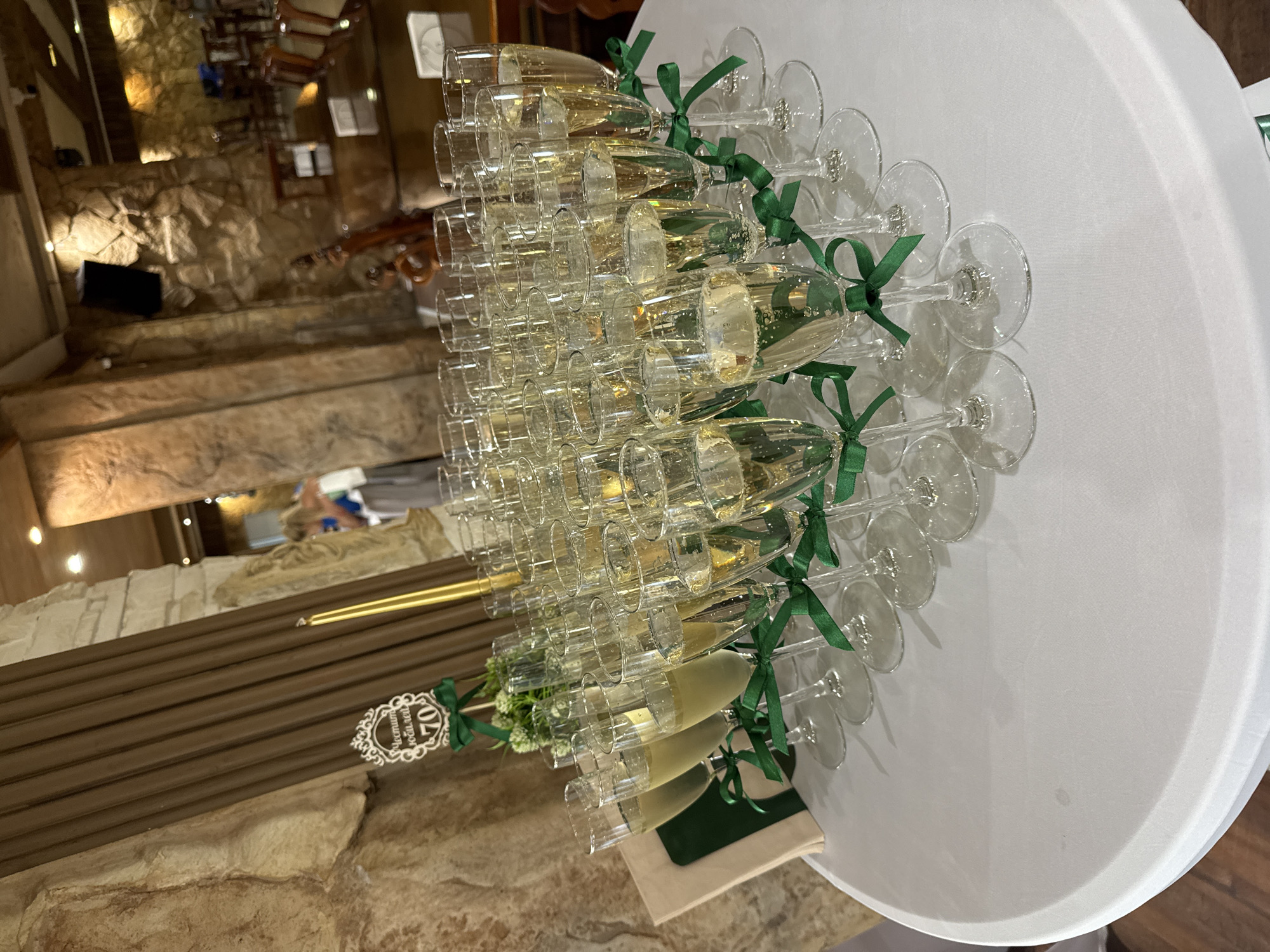 A circular white tablecloth is spread across a table, with many glasses of champagne arranged in rows. The glasses are tall and slender, filled with a light yellow bubbly liquid, and each is adorned with a green ribbon tied around its stem. A decorative sign with the number "70" is visible on the left side of the table, along with a small bouquet of green foliage. Behind the table, a dimly lit interior with stone walls and wooden paneling is visible. In the background, several people are blurred and out of focus. The scene appears to be set at an event or celebration. The location is the city of Sredets, Bulgaria.