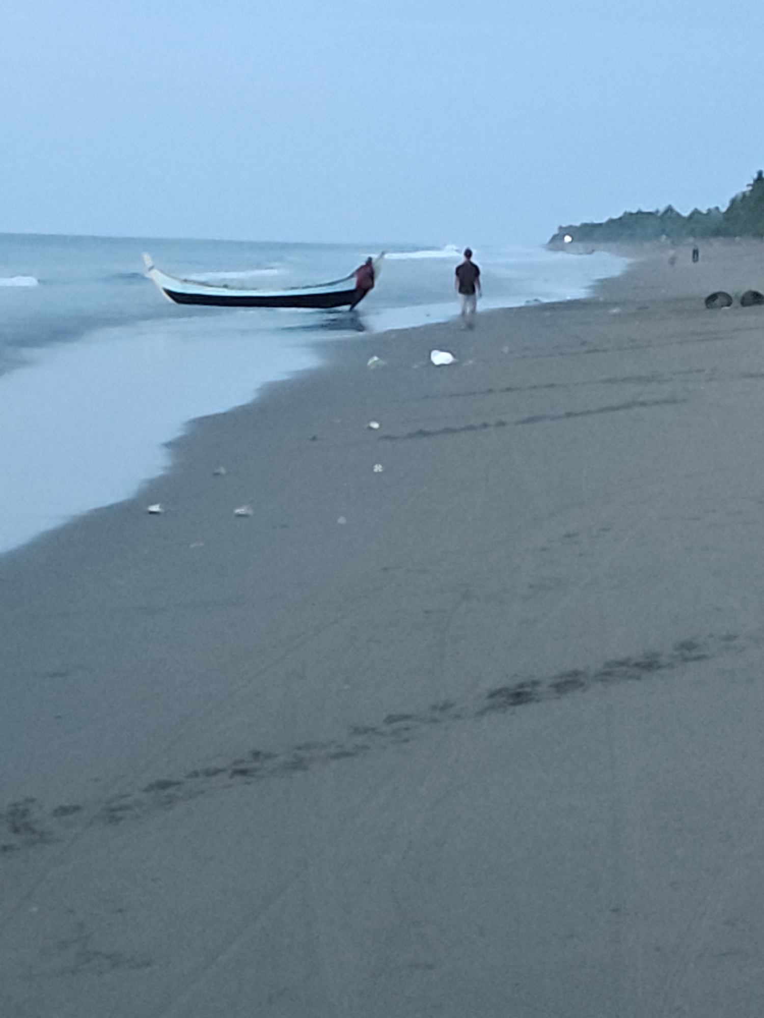 The image depicts a tranquil scene on a beach in Curehtunong, Indonesia, during what appears to be either dawn or dusk, as indicated by the soft, diffused light. A traditional Indonesian fishing boat, with its distinctive curved hull and outriggers, rests partially on the wet sand at the water's edge. The boat is dark in color with a lighter-colored hull. A figure, wearing a red garment and dark pants, is positioned near the stern of the boat, suggesting they may be tending to it or preparing for a voyage.
Further down the beach, a single individual, dressed in a dark t-shirt and light-colored pants, is walking away from the water and towards the interior of the beach. Their posture indicates a casual stroll. The beach itself is composed of fine, light-colored sand. In the distance, a line of lush green trees forms a dense backdrop, with a hint of distant lights suggesting a settlement or activity beyond the immediate coastline. The ocean is calm, with gentle waves lapping at the shore. The overall atmosphere is serene and peaceful, conveying a sense of everyday life in this coastal Indonesian community.