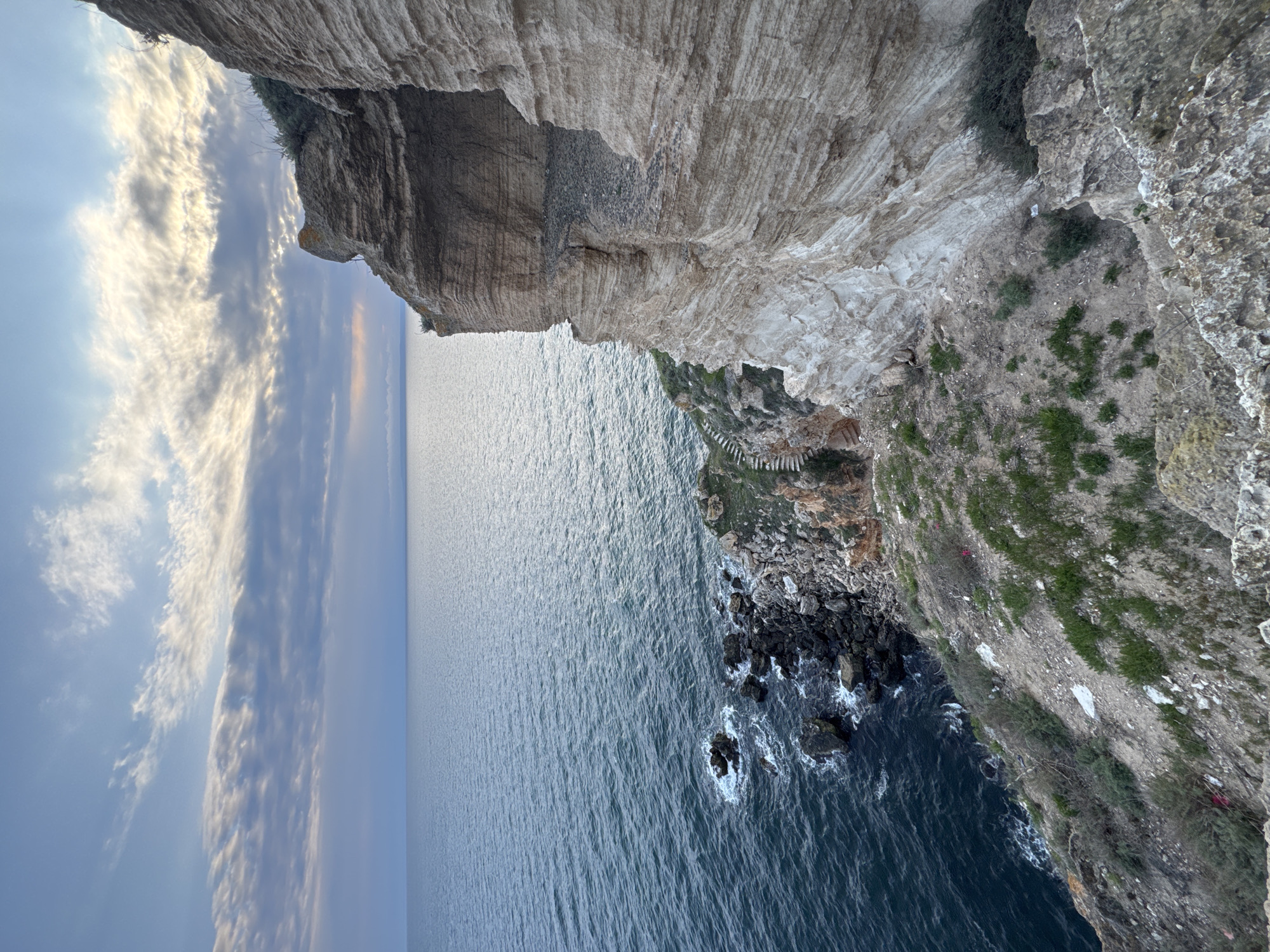 The image depicts a cliffside overlooking the Black Sea near Balgarevo, Bulgaria. The cliff face shows stratified rock formations, with some vegetation growing on its slopes. At the base of the cliff, rocks are visible where the cliff meets the sea, with waves crashing against them. In the distance, the sea stretches to the horizon where it meets a sky with cloud formations. A set of stairs is built into the cliffside.