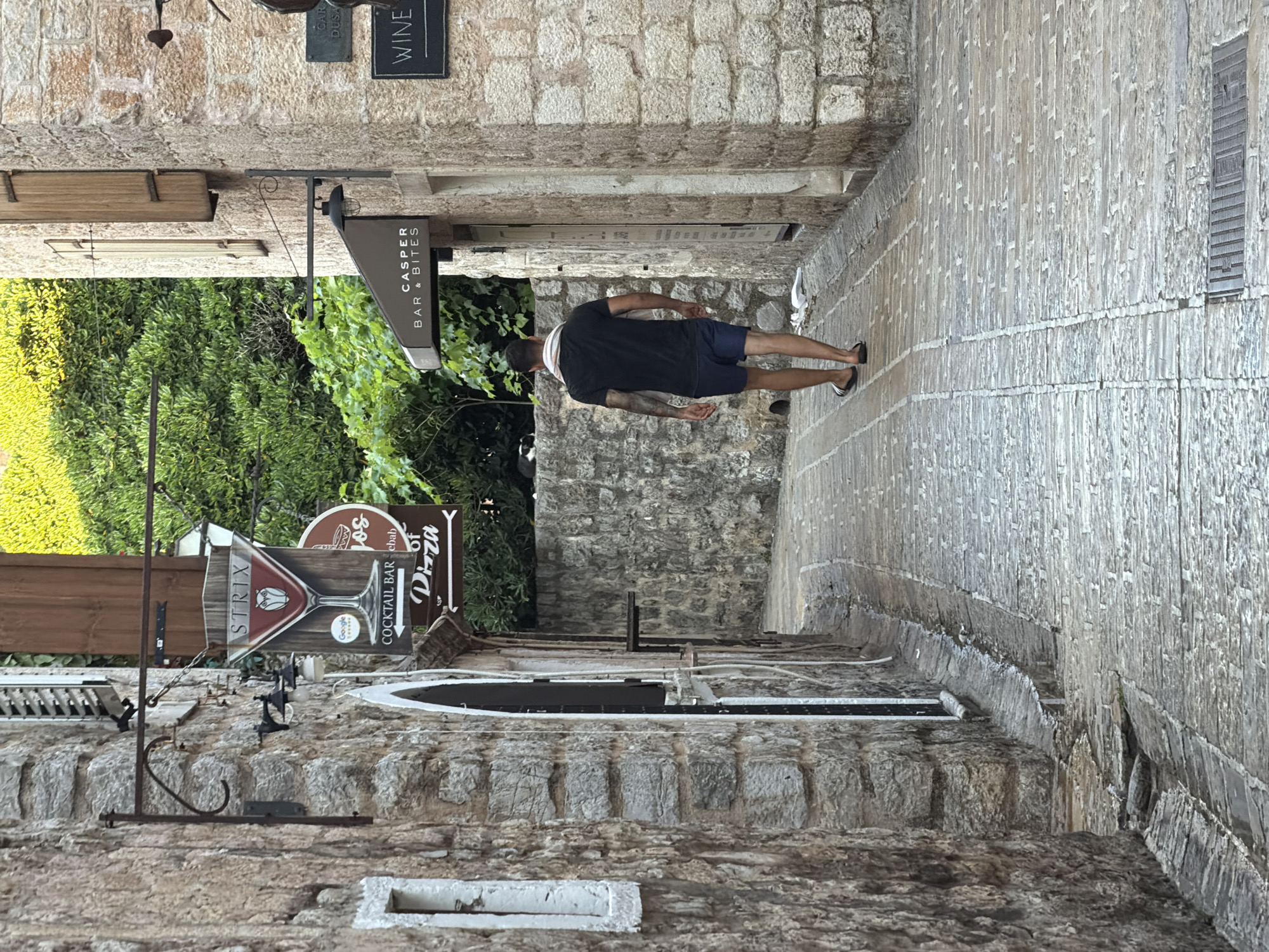 A man walks down a cobblestone alleyway in the Old Town of Budva, Montenegro. The man, wearing a black t-shirt, blue shorts, and flip-flops, has a towel slung around his neck and is facing away from the camera. He is walking towards a street with a sign that reads "STRIX COCKTAIL BAR Kebab of Pizza" on the left side of the alley, and a sign that reads "CASPER BAR & BITES" on the right side of the alley. Further down the alley on the right, a sign reads "WINE". The buildings flanking the alley are constructed of rough stone. Lush green foliage can be seen in the background. The alley appears to be somewhat damp, with a drain running along the right side of the street.