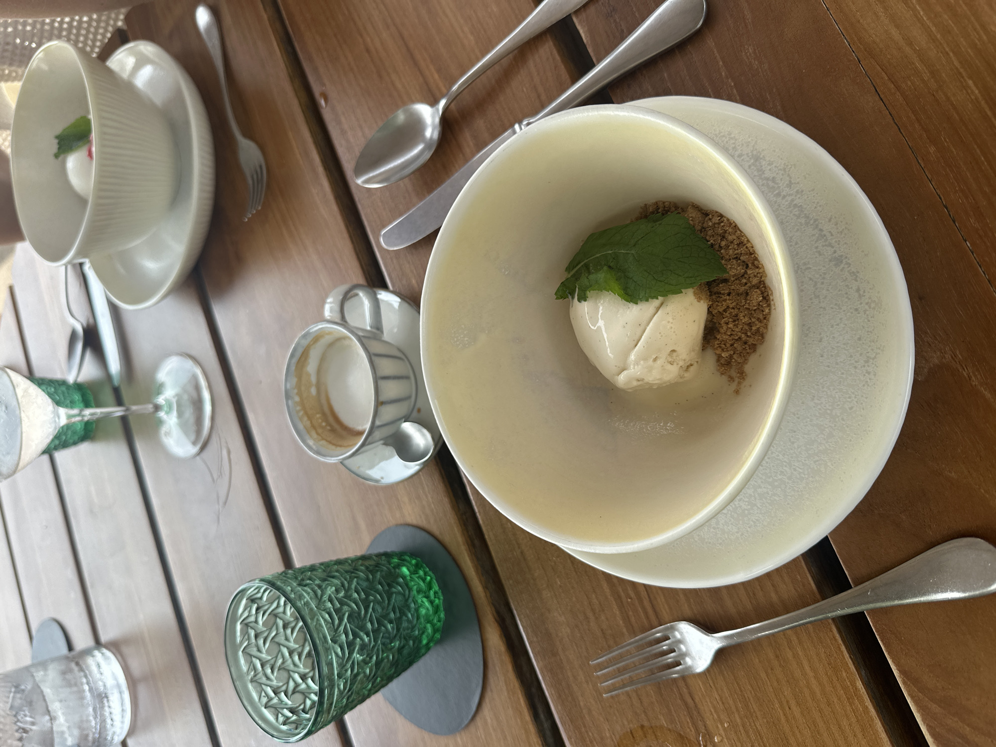 An overhead, medium shot captures a dining table set for one, likely outdoors given the wooden planks. The table is adorned with a dessert in a white bowl, two glasses (one green, one clear with water), a cup of coffee, and cutlery. The dessert, a scoop of light-colored ice cream with a sprig of mint and crumbled cookie base, is positioned prominently in the foreground. To the left, a textured green glass, likely holding a beverage, sits on a dark coaster. Adjacent to it, a clear glass with water is visible. In the background, a wine glass with a light-colored liquid and a delicate white bowl containing a small pink item and a mint leaf are present. A small demitasse cup filled with coffee rests on a saucer with a spoon. Cutlery, including a fork, knife, and spoon, is strategically placed around the dishes. The wooden surface of the table shows a subtle grain and appears slightly damp in places, suggesting recent use or a humid environment. The scene is lit by natural, soft light, casting gentle shadows. No people are visible in the image.