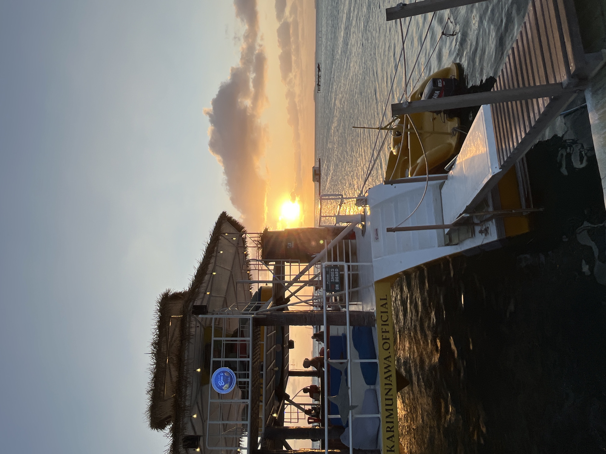 The image depicts a sunset scene at a waterfront establishment in Karimunjawa, Indonesia. The establishment, likely a restaurant or bar, features a two-story structure with a thatched roof and open-air seating areas. A sign with the words "KARIMUNJAWA.OFFICIAL" is visible on the lower level, indicating its location. Several people are seated at tables on the upper level, and beanbag chairs are arranged on the lower level. A yellow boat with an outboard motor is docked beside the structure. A wooden ramp extends from the establishment to the water. The sun is setting over the ocean, casting a warm glow on the scene. The water is calm, with reflections of the setting sun visible on its surface. The sky is a gradient of blue and orange, with scattered clouds. The overall atmosphere is serene and tranquil.
