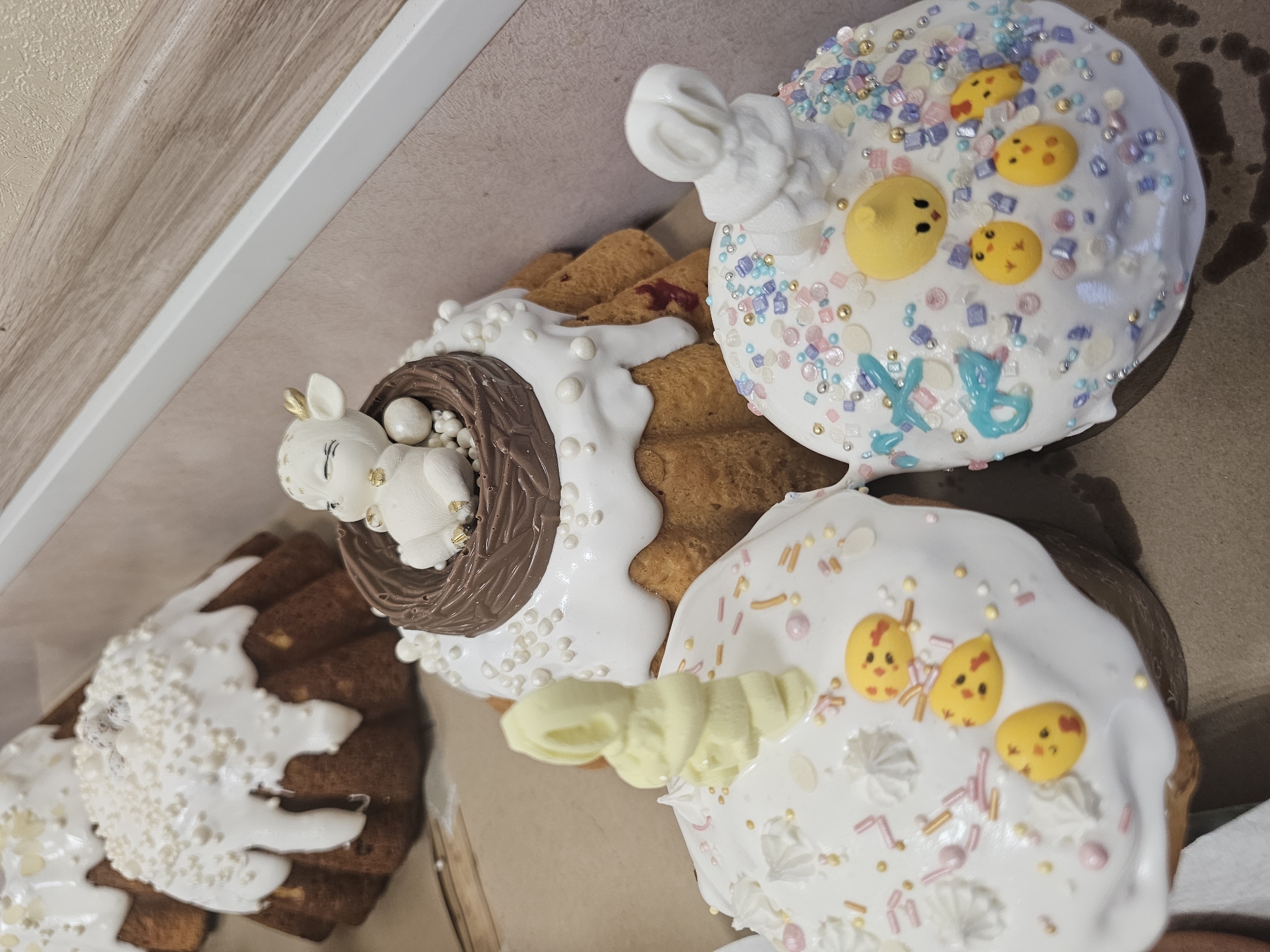 The image shows a close-up, overhead view of several elaborately decorated Easter cakes, likely inside a box. The cakes have white frosting and various decorative elements.
One cake is topped with a small, sculpted rabbit lying in a chocolate nest, adorned with white pearl-like decorations. Other cakes have decorations including small yellow chick figures and colorful sprinkles. Some cakes are decorated with swirls of white frosting. One cake has blue Cyrillic letters that appear to say "XB" which is a common abbreviation for "Христос Воскресе" (Christ is Risen), a traditional Easter greeting.
The cakes appear to be commercially produced, possibly for sale in Moscow, Russia given the Easter theme and the Cyrillic inscription.