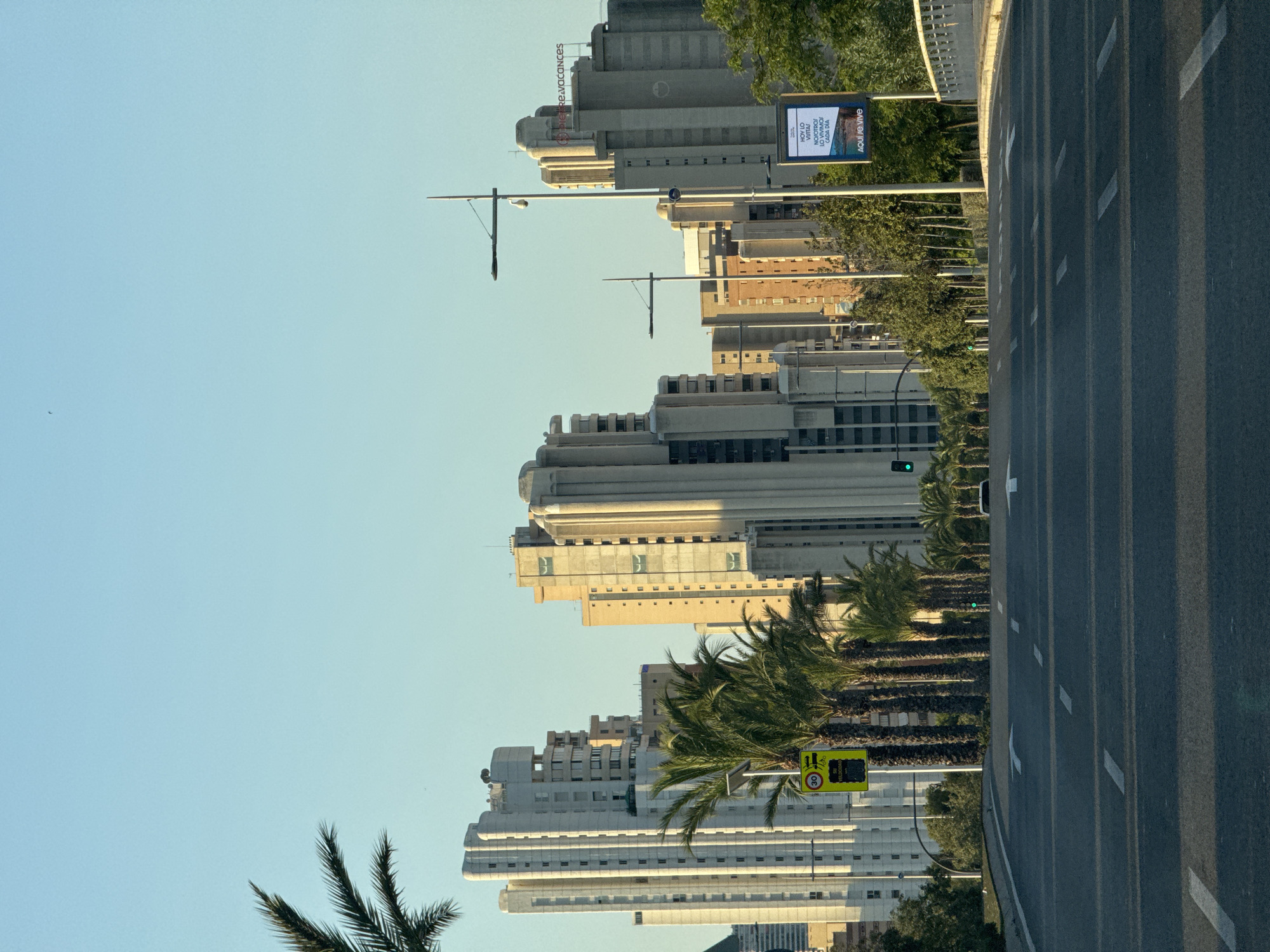 The image shows a wide, paved road leading towards a city skyline dominated by tall, modern buildings. The architecture of the buildings suggests a coastal resort city. Palm trees are interspersed along the sides of the road and in front of the buildings, indicating a warm climate. On the left side of the road, a traffic sign displaying "30" and symbols for a road and a bicycle is visible. Several traffic lights are also present, some showing green. A billboard with Spanish text, reading "HOY LO VISITAS! NOSOTROS LO VIVIMOS CADA DIA. AQUI SE VIVE", is positioned on the right side of the road, near some greenery. The sky is clear and blue, with the sun casting long shadows, suggesting either early morning or late afternoon. The scene appears to be in Benidorm, Spain, a city known for its high-rise buildings and beach resorts.