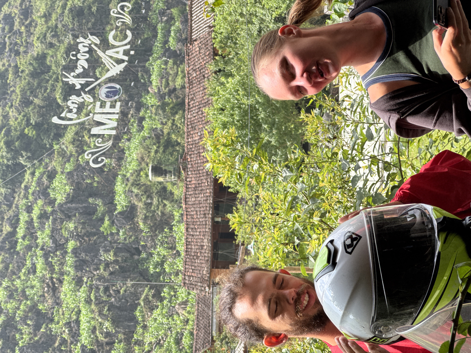 Two people pose for a photo in Mèo Vạc Commune, Vietnam. The woman, with a ponytail and olive shirt, holds a phone. The man with a beard is holding a motorcycle helmet. Behind them is a lush green mountain, the roof of a building, and various vegetation. The location sign "Cung Đường Mèo Vạc" can be seen on the mountain.
