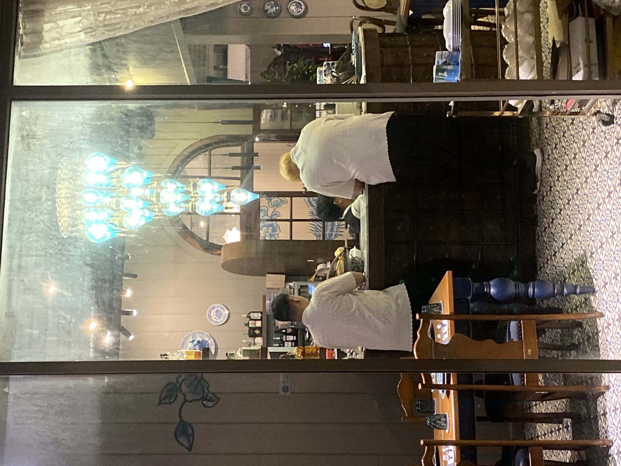 A scene viewed through a glass storefront shows two people at a counter, presumably within a commercial establishment in Choeng Thale, Thailand. One person, with light blonde hair, is wearing a white shirt and dark pants, and is bent over the counter. Another person is standing and wearing a white shirt, also appears to be engaged with the counter. Various bottles, possibly containing alcohol or beverages, line shelves behind the counter. The storefront's window reflects an interior chandelier composed of blue bulbs and other decorative elements, creating a layered perspective. Furnishings inside include wooden chairs.