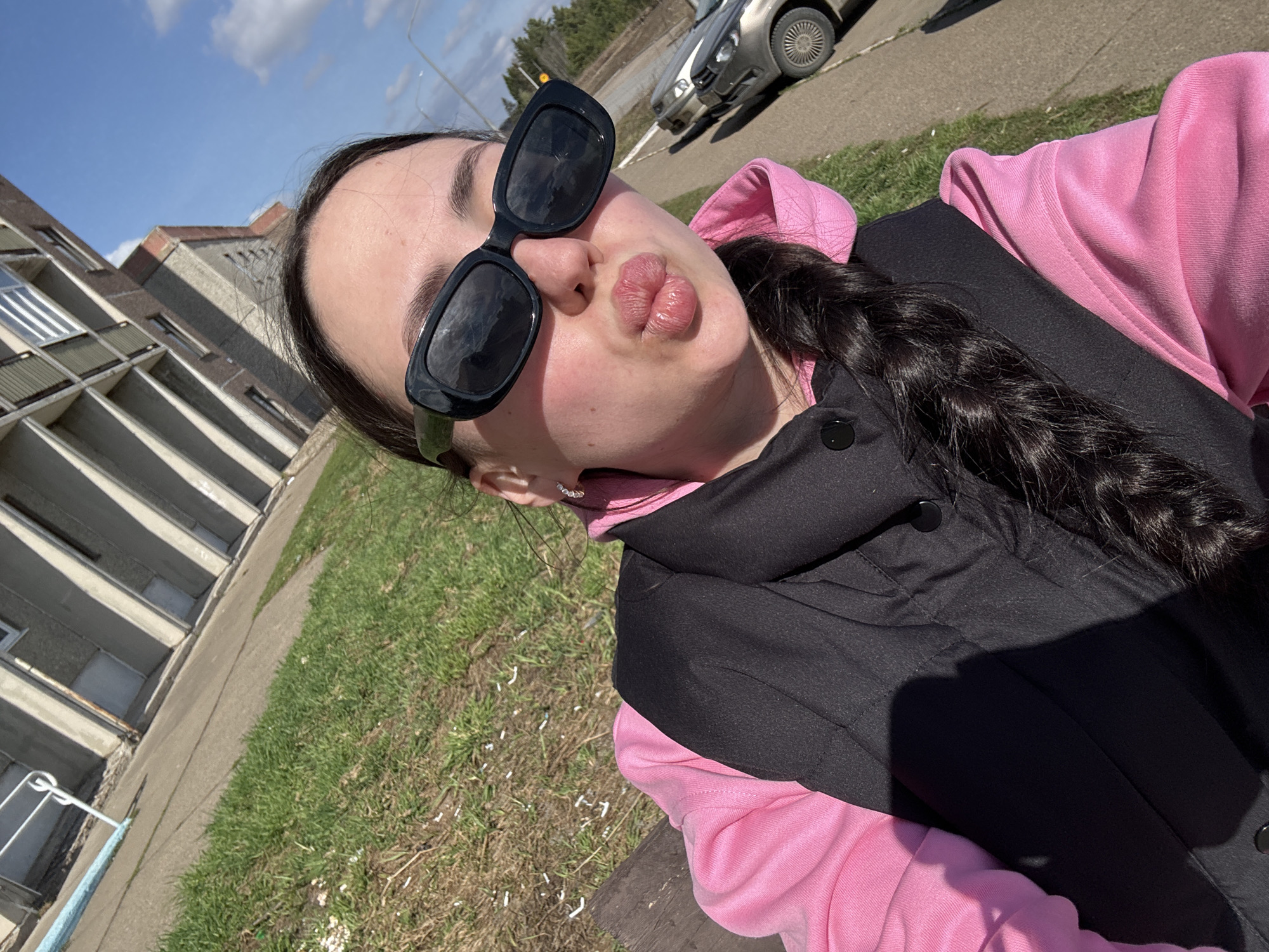 A close-up, angled selfie features a person, appearing female, on a bright, sunny day. The subject is wearing large black rectangular sunglasses, a vibrant pink hooded sweatshirt, and a black puffer vest. Her dark hair is styled in a braid, visible over her left shoulder. She is making a playful "kissing" face with puckered lips.
The outdoor setting includes a multi-story concrete building with numerous windows and balconies in the background, suggesting an apartment block or utilitarian structure. A paved area or sidewalk is visible, alongside a patch of green grass scattered with small white debris, possibly petals or paper. A light-colored car is parked on a street further back. The sky is clear blue with some scattered white clouds, and strong shadows indicate bright sunlight, suggesting it is likely late morning or afternoon. The overall atmosphere is casual and cheerful. No visible text is present in the image.