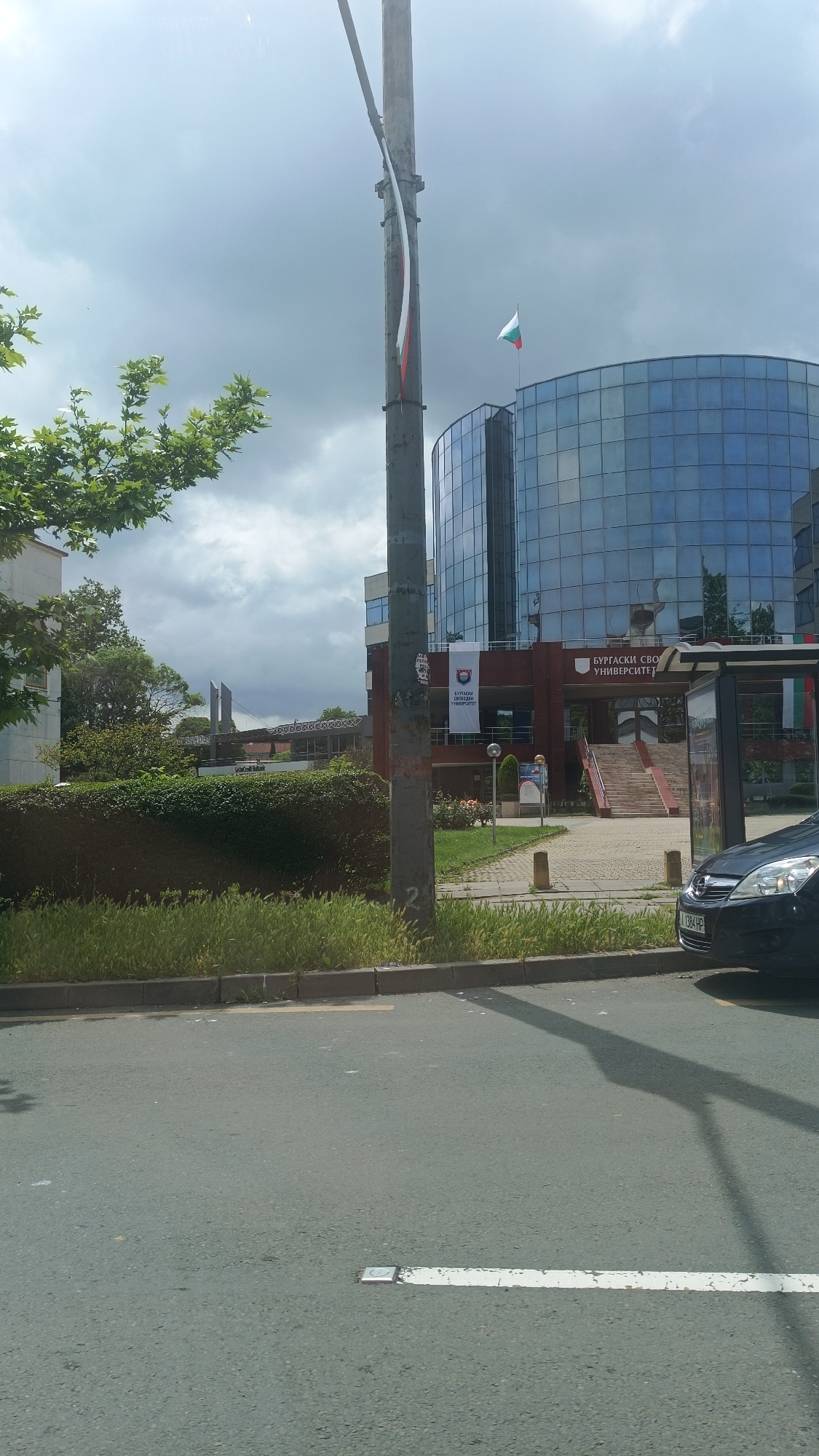 The image depicts a street scene in Burgas, Bulgaria, featuring Burgas Free University. The university's distinctive architecture includes a rounded, glass-clad building visible in the background. A Bulgarian flag flies atop this building. A bus stop is located on the right side of the frame, partially obscured by a dark-colored car. A lamp post stands in the foreground, with what appears to be a tattered or partially unfurled Bulgarian flag attached to it. The street is marked with a white painted line. Green shrubbery separates the street from the university grounds. The sky is overcast.