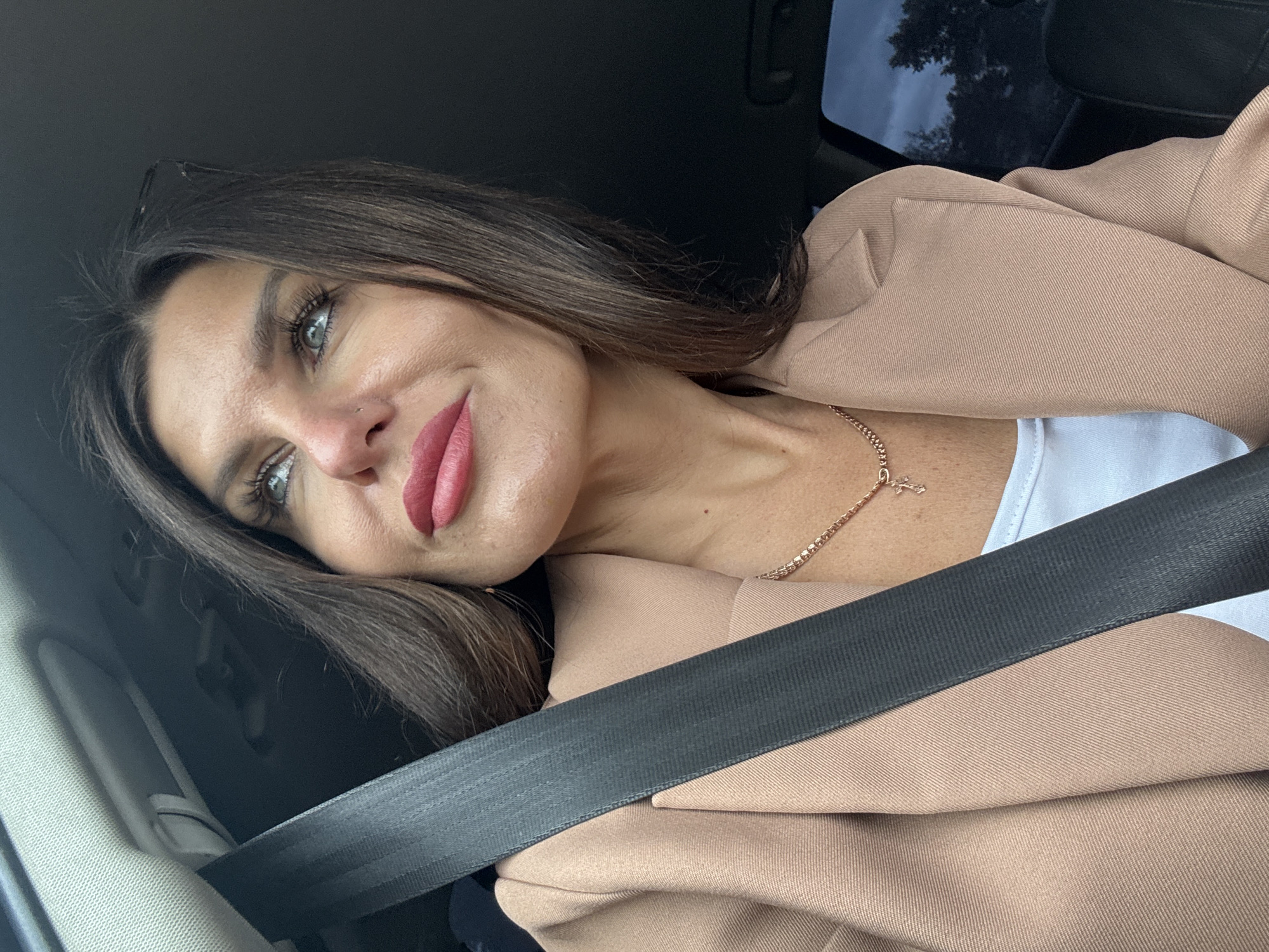 A woman with long brown hair and green eyes is in the driver's seat of a car. She is wearing a light beige blazer over a white top and a delicate gold necklace with a cross pendant. A black seatbelt is fastened across her chest. Her makeup includes bold pink lipstick and mascara. She is looking to her left, with a slight smile and a thoughtful expression. The background shows the interior of a car, including a dark grey dashboard and the reflection of trees and a cloudy sky in the window. The lighting suggests it is daytime, possibly overcast, given the dim interior and the sky's appearance. The location is identified as Chaikovsky, Russia.