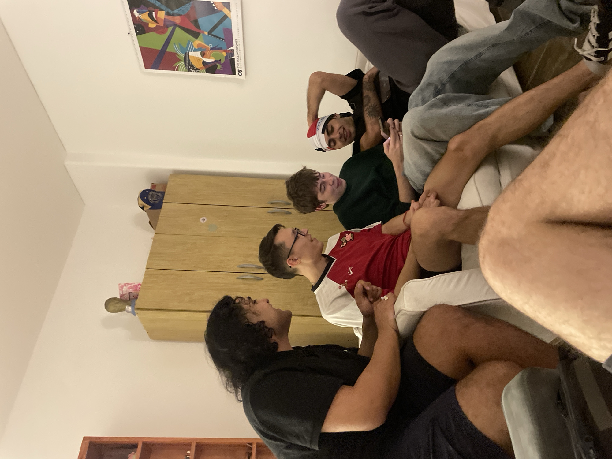 An indoor scene depicts a casual gathering of several young men in what appears to be a living room in Buenos Aires, Argentina. The room has plain white walls and wooden furniture, including a cabinet and shelves.
Multiple individuals are visible, mostly seated on or around a light-colored couch. One man, smiling broadly, wears a black t-shirt and a red and white Santa-style headband. Another man in a dark green long-sleeved shirt is partially visible, looking downwards. A third man, wearing glasses and a red and white short-sleeved shirt (possibly a soccer jersey), looks upwards. To the foreground, a man with long dark hair in a black t-shirt appears to be speaking, looking upwards and to the left. Legs and parts of other individuals are also visible, suggesting more people are present but not fully in frame.
The group seems to be socializing and engaging in conversation. The festive headband suggests a playful or holiday-themed gathering.
Notable details include a colorful poster on the wall with the text "THE ROLLING STONES" and "05" visible. The room is well-lit, but the time of day is not explicitly indicated by natural light. Emotions appear relaxed and engaged, with one clear smile. No specific weather cues are present as it's an indoor scene.