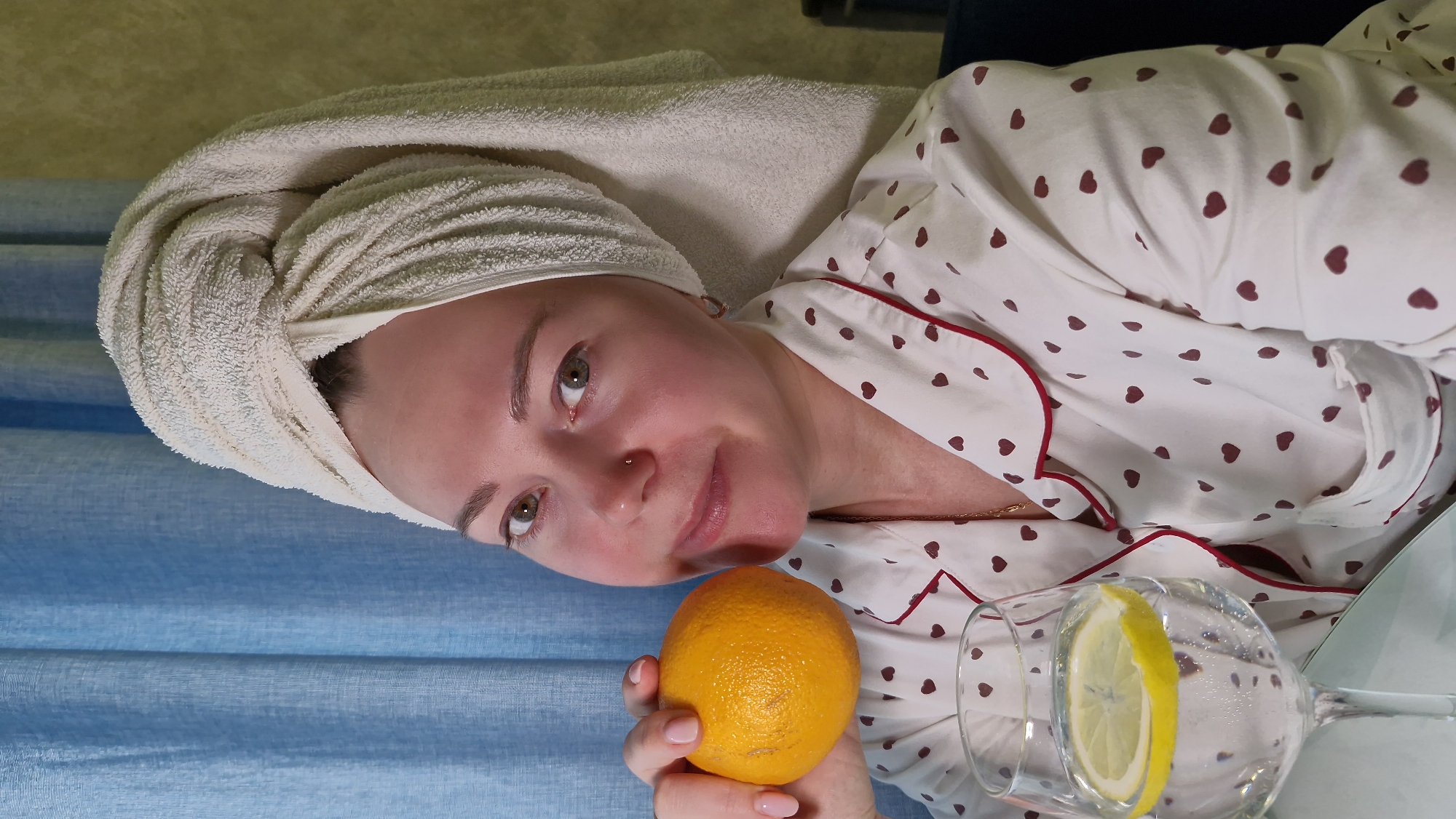 A woman with a towel wrapped around her head holds an orange and a glass of water with a lemon slice. She wears a white pajama top with red heart-shaped patterns and red trim. A light blue curtain is visible in the background. The city of Otchor, Russia, cannot be determined from the image.