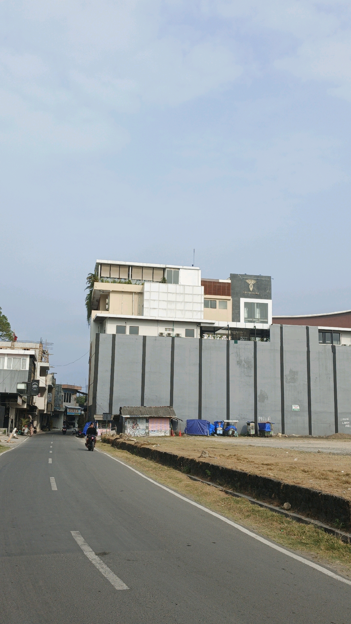 In Desa Pagandaran, Indonesia, a road curves through a semi-urban environment under a pale sky. On the right side of the road, a large, multi-story building with a gray facade dominates the view. The building has a modern design with balconies and windows. In front of this building, there are several small stalls or temporary structures, some covered with blue tarps, and two small, covered vehicles, possibly rickshaws or small carts. A motorcycle rider is visible on the road in the mid-ground, heading away from the viewer. On the left side of the road, a row of smaller, older buildings lines the street, some with visible signage, including one that reads "SURYA." The area appears to be a mix of commercial and residential development, with some areas of undeveloped land with dry grass along the roadside. The overall impression is one of a functional, developing area.