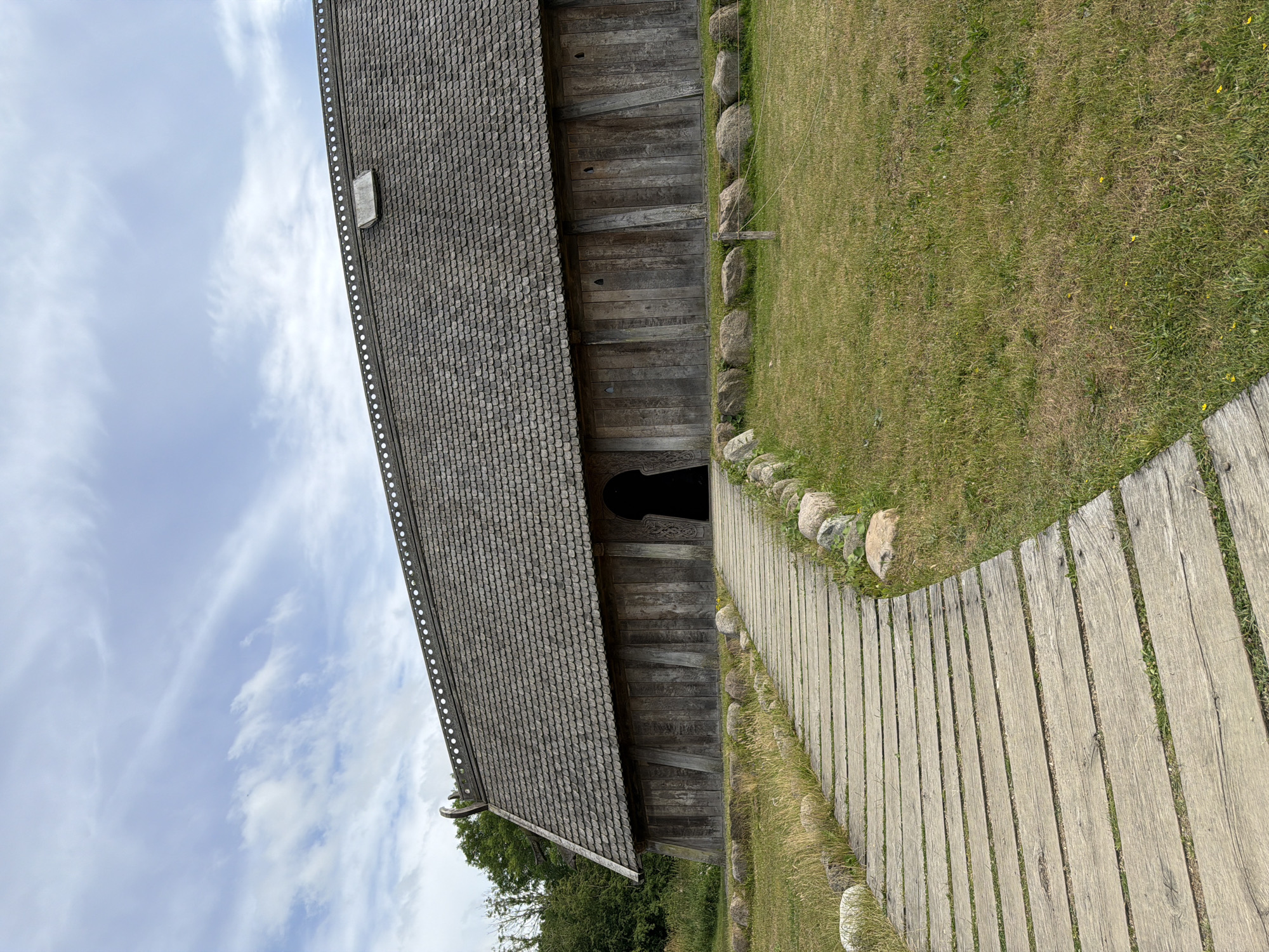 The image shows an outdoor scene of a historical building with a wooden plank walkway leading to its entrance. The building has a steeply pitched roof covered in wooden shingles and a dark, arched entranceway. The walkway is made of weathered wooden planks laid side by side, curving slightly to the right. Large, rounded stones are placed along the edge of the walkway, separating it from a grassy area. The grass appears somewhat dry in places, with patches of green and yellow. A few small yellow wildflowers are scattered in the grass.
The sky is a pale blue with wispy white clouds. The lighting suggests it is daytime, possibly late morning or afternoon, with soft, diffused light. The weather appears to be fair and possibly a bit overcast.
To the left of the building, there is a glimpse of green trees, suggesting a natural or park-like setting. The overall scene evokes a sense of history and rustic charm. The building's architecture suggests it could be a replica of a Viking longhouse or a similar historical structure, consistent with the location context of Allerslev, Denmark, known for its historical sites. There is no visible text in the image. No people are visible in the scene, and the focus is on the building, the walkway, and the surrounding natural elements.
