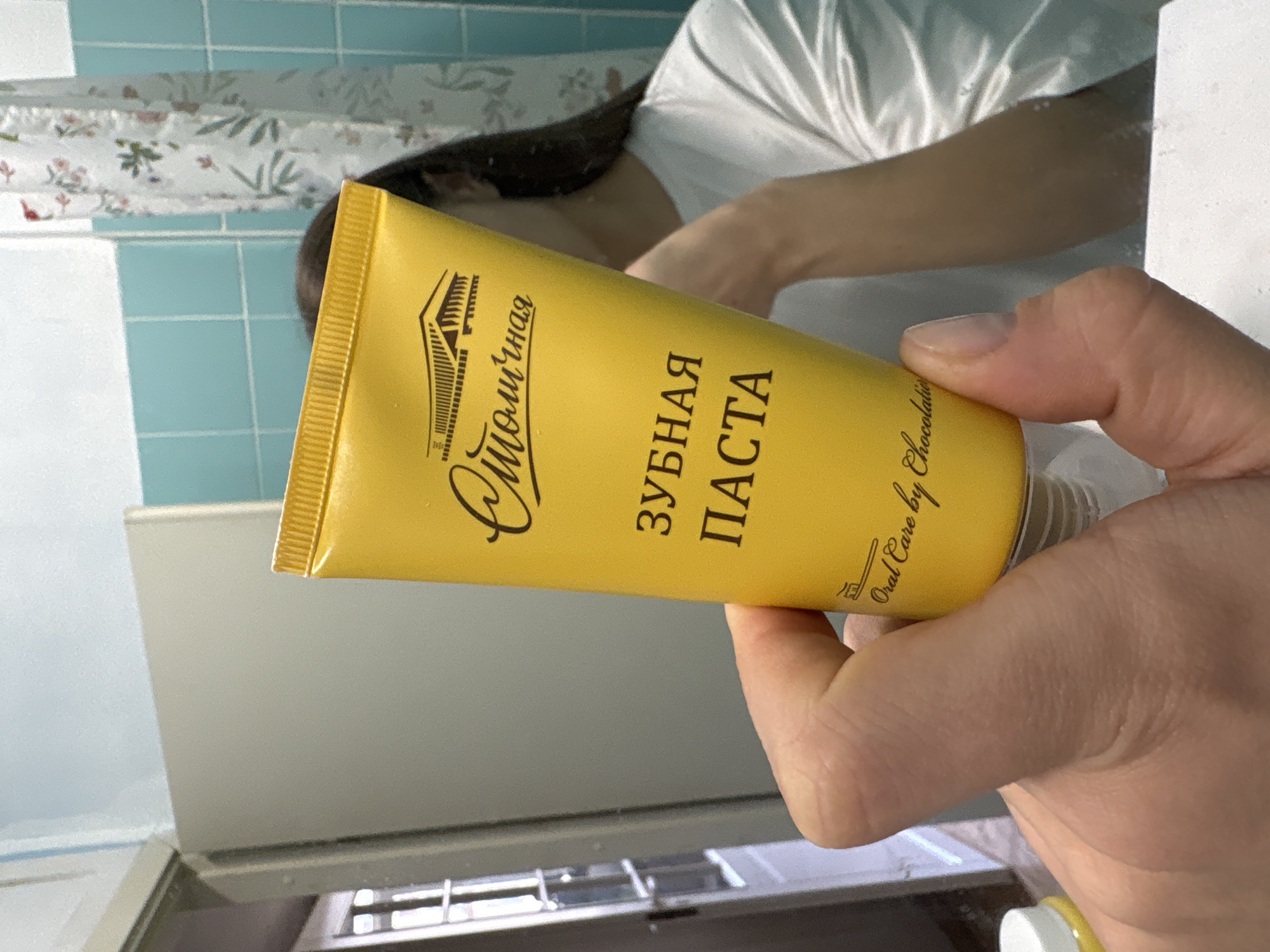 A hand holds a yellow tube of "Oral Care by Chocolate" toothpaste, labeled in Russian as "ЗУБНАЯ ПАСТА" and "Омолочная" which is also the name of the manufacturer. The background includes a reflection of a person wearing a white shirt. The immediate background shows a bathroom with turquoise tiles and a shower curtain with a floral pattern. A white sink is also visible. There is no verifiable evidence from the image to definitively state that the location is Moscow, Russia.
